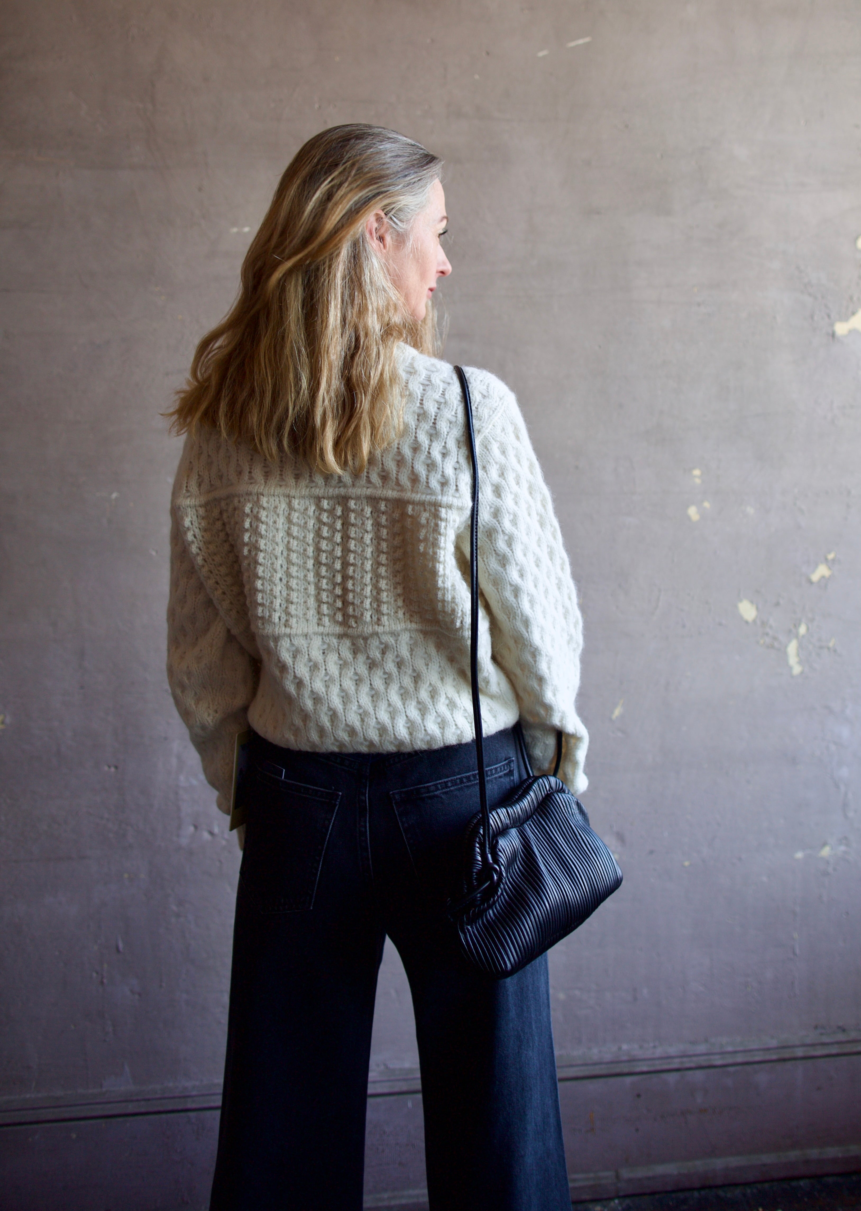 Image featuring a woman wearing the Baci handbag by Mansur Gavriel in black with a magnetic closure, adjustable crossbody strap and intricate pintucked leather across entire hand bag.