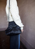 Image featuring a woman wearing the Baci handbag by Mansur Gavriel in black with a magnetic closure, adjustable crossbody strap and intricate pintucked leather across entire hand bag.