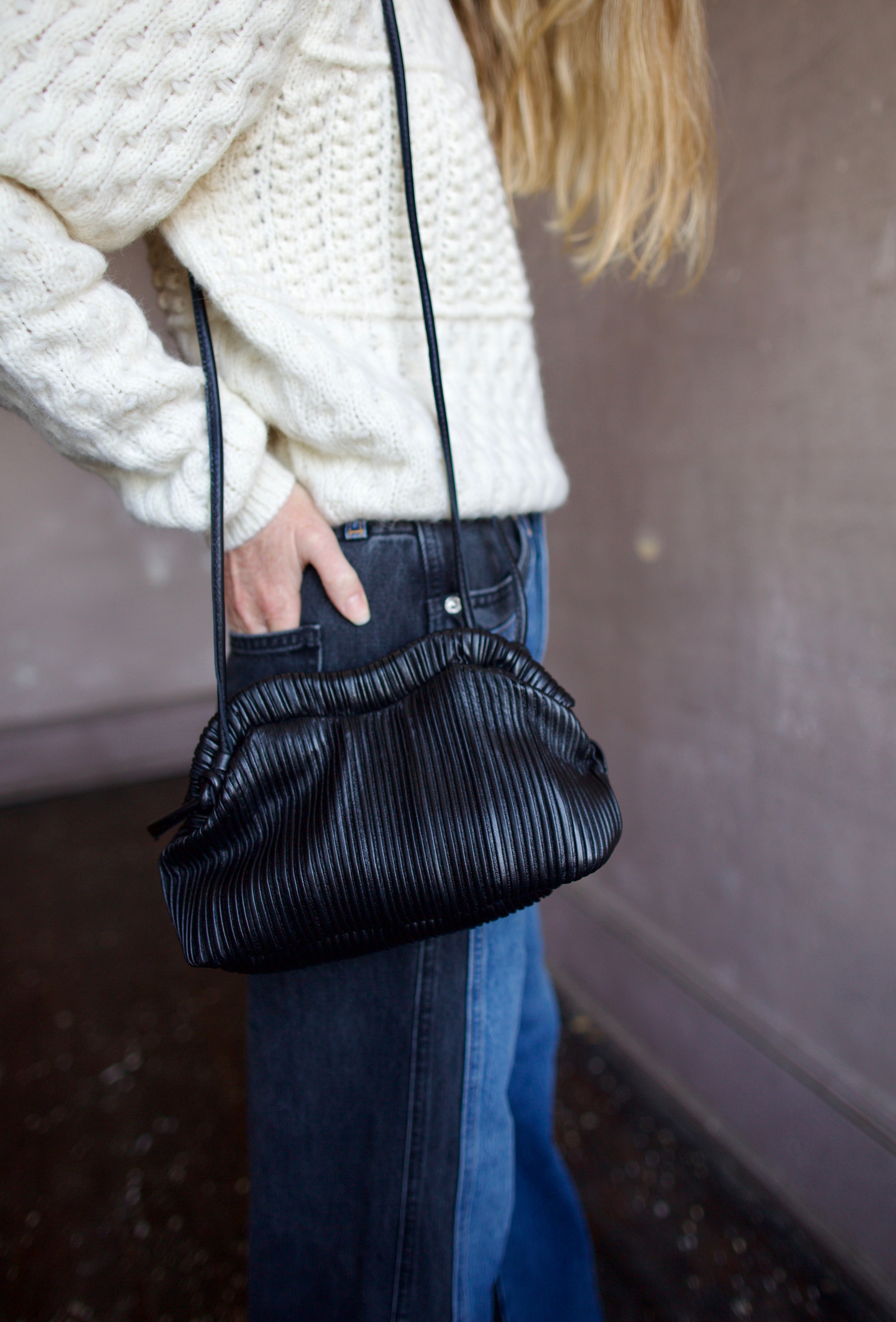 Image featuring a woman wearing the Baci handbag by Mansur Gavriel in black with a magnetic closure, adjustable crossbody strap and intricate pintucked leather across entire hand bag.