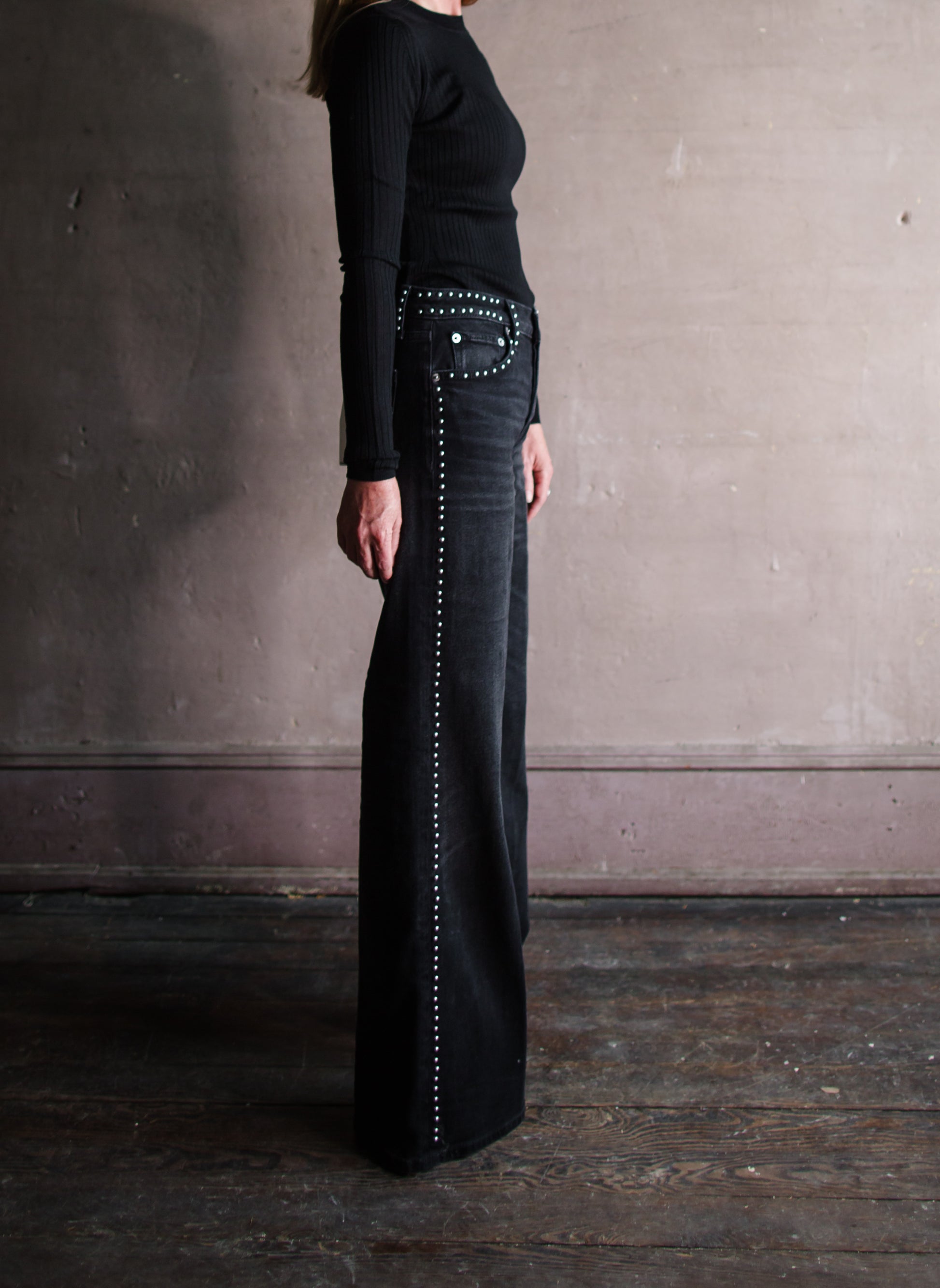 Image featuring a woman wearing the black jeans by Re/Done with a mid rise and wide leg embellished with silver studs down leg seams, outlining pockets and waistband.