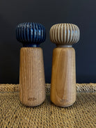 A wooden spice grinder set with a neutral taupe beige and navy blue ribbed ceramic top and a natural finish oak wood body