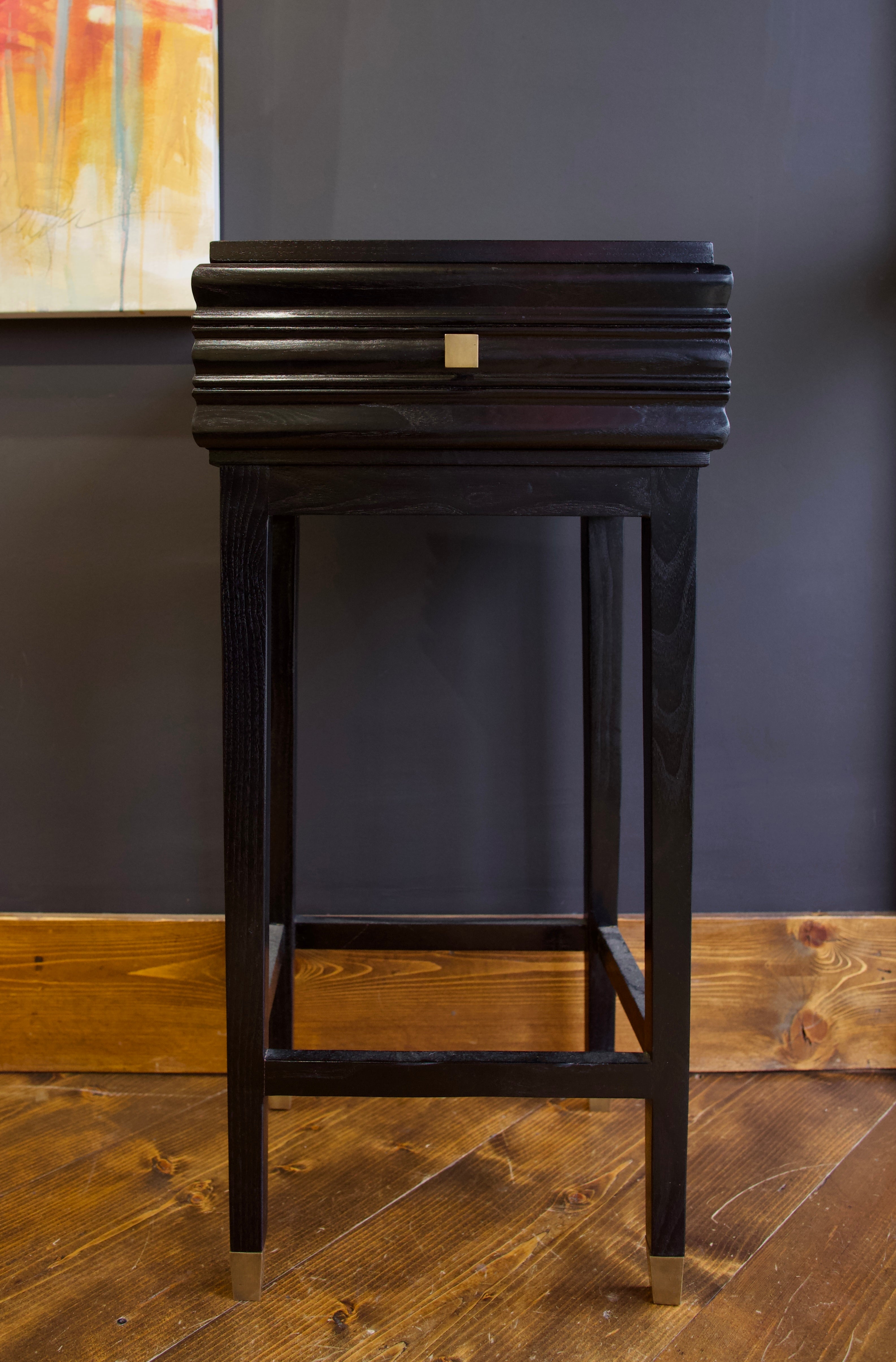 A tall black side table with a single contoured drawer, square legs and brass hardware