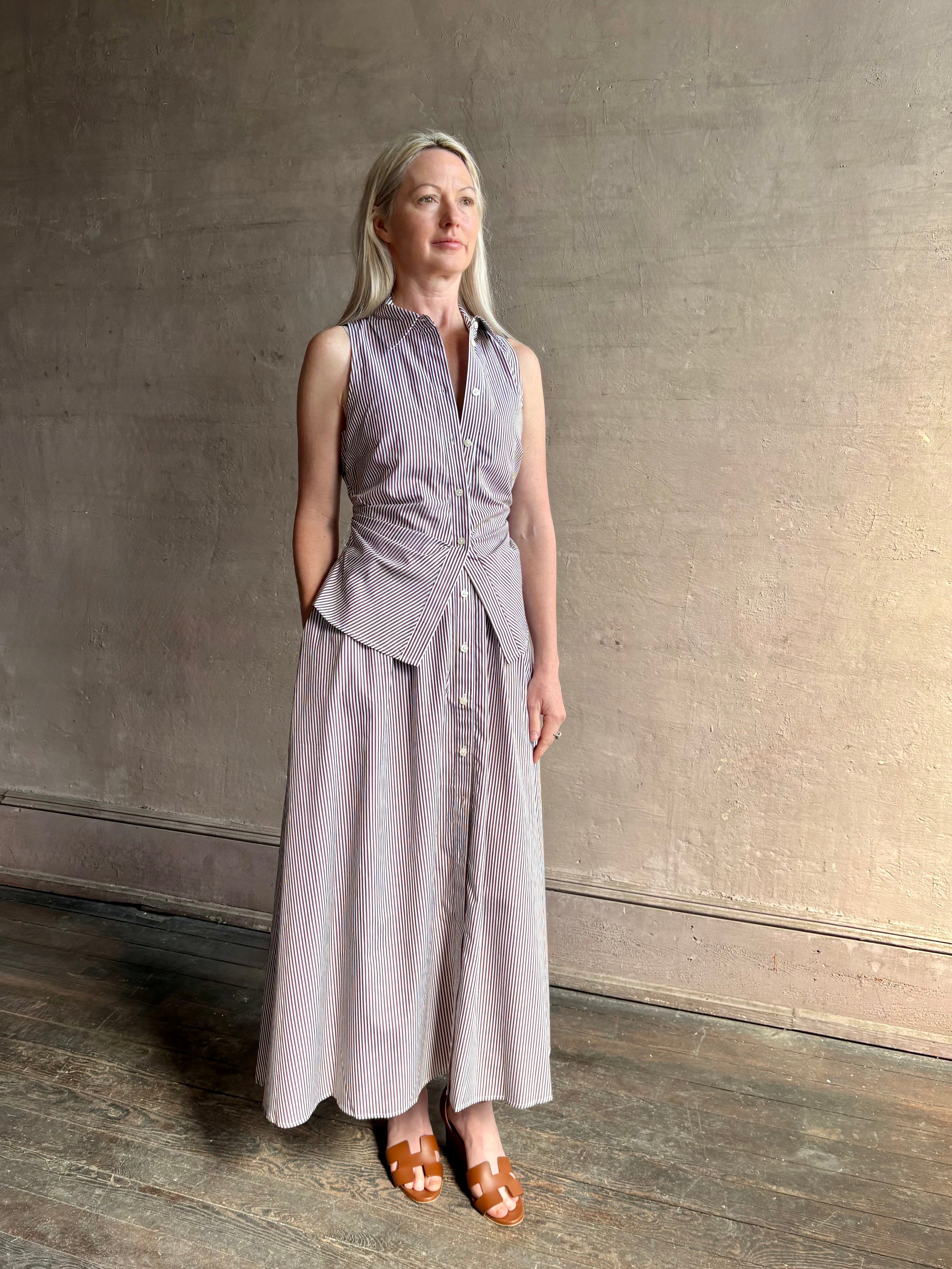 Mature woman wearing the Hannah maxi shirt dress from designer Tanya Taylor rendered in a brown and white stripe and crafted from 100% cotton poplin with a very classy flattering waistline that doesn't stick to body, sleeveless with a collar for a timeless tailored classic dress