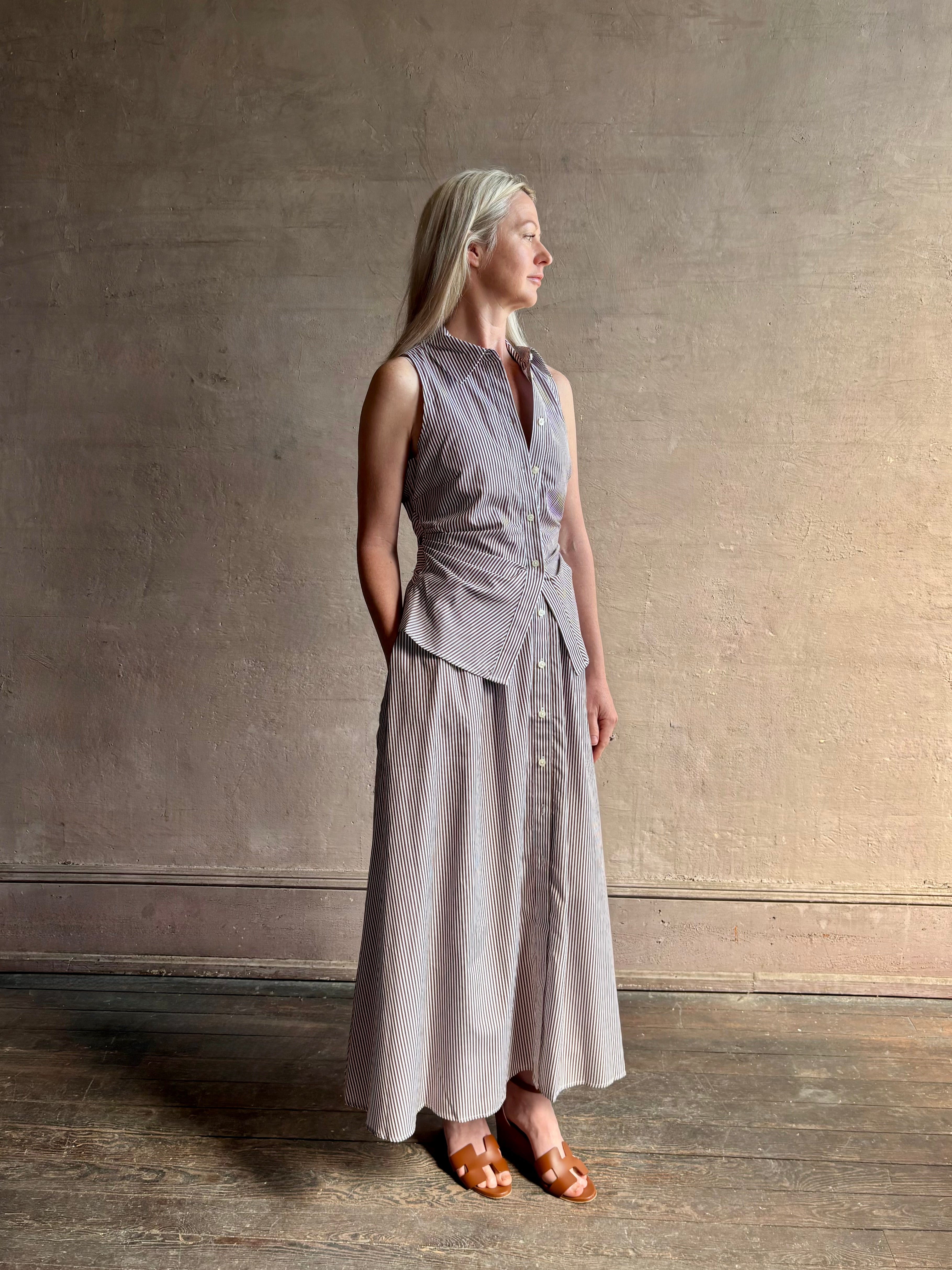 Mature woman wearing the Hannah maxi shirt dress from designer Tanya Taylor rendered in a brown and white stripe and crafted from 100% cotton poplin with a very classy flattering waistline that doesn't stick to body, sleeveless with a collar for a timeless tailored classic dress