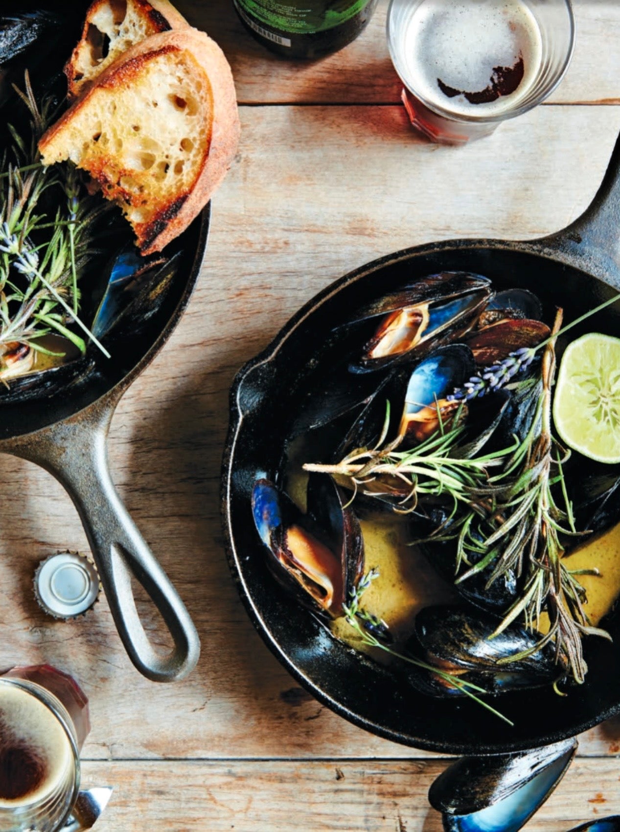 Cast iron skillet with mussels on a rustic wooden table, garnished with rosemary and a lime wedge, alongside toast 
