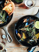 Cast iron skillet with mussels on a rustic wooden table, garnished with rosemary and a lime wedge, alongside toast 