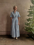 Mature woman wearing Martine maxi shirt Dress from Trovata rendered in a blue and white Bahia Stripe and crafted from cotton poplin with ruffle lined short sleeves, collar, side seam pockets, button front, tiered skirt and optional self tie co-ordinating fabric belt
