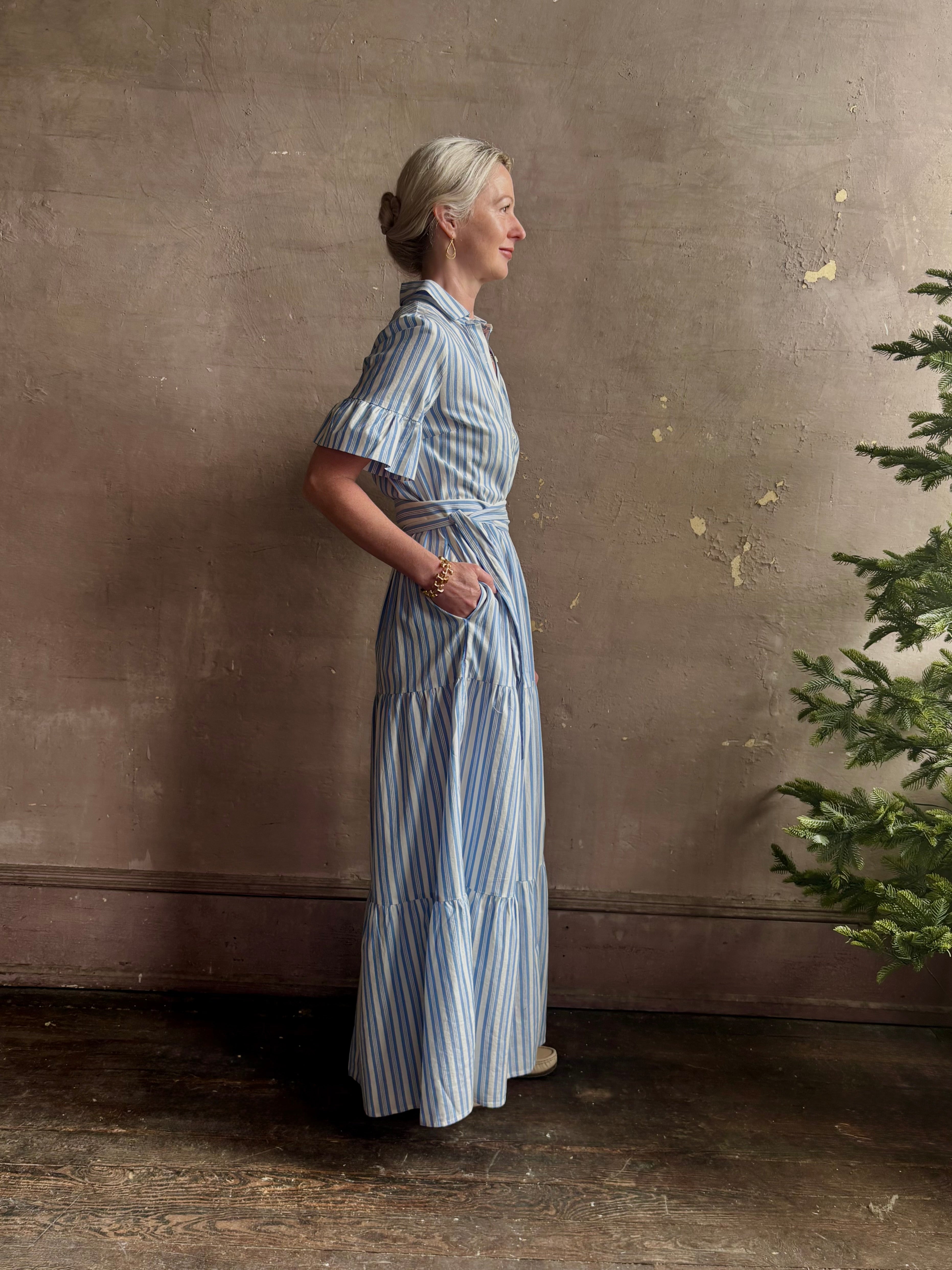 Mature woman wearing Martine maxi shirt Dress from Trovata rendered in a blue and white Bahia Stripe and crafted from cotton poplin with ruffle lined short sleeves, collar, side seam pockets, button front, tiered skirt and optional self tie co-ordinating fabric belt