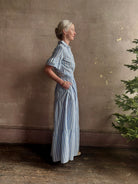 Mature woman wearing Martine maxi shirt Dress from Trovata rendered in a blue and white Bahia Stripe and crafted from cotton poplin with ruffle lined short sleeves, collar, side seam pockets, button front, tiered skirt and optional self tie co-ordinating fabric belt