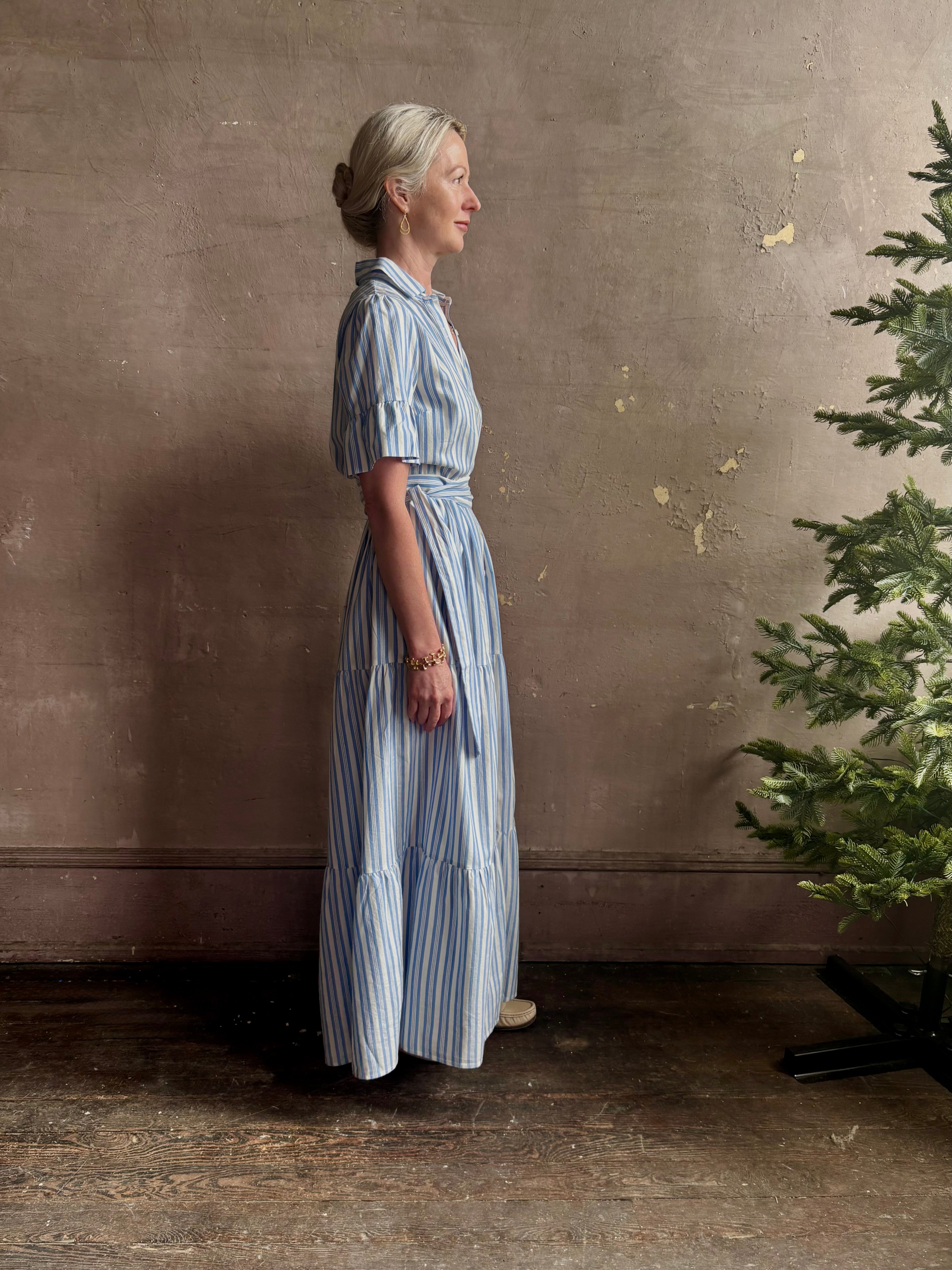 Mature woman wearing Martine maxi shirt Dress from Trovata rendered in a blue and white Bahia Stripe and crafted from cotton poplin with ruffle lined short sleeves, collar, side seam pockets, button front, tiered skirt and optional self tie co-ordinating fabric belt