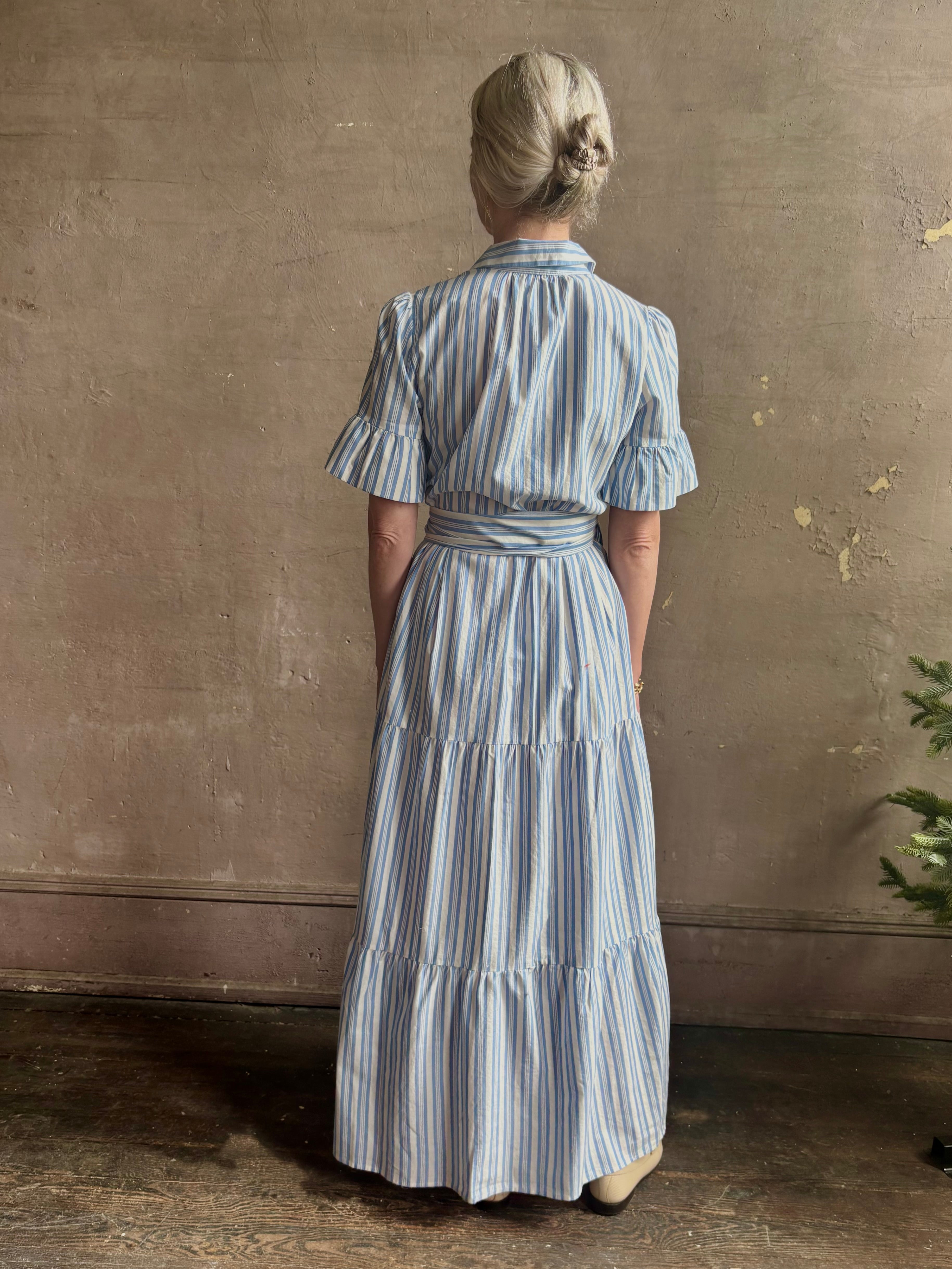 Mature woman wearing Martine maxi shirt Dress from Trovata rendered in a blue and white Bahia Stripe and crafted from cotton poplin with ruffle lined short sleeves, collar, side seam pockets, button front, tiered skirt and optional self tie co-ordinating fabric belt