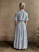 Mature woman wearing Martine maxi shirt Dress from Trovata rendered in a blue and white Bahia Stripe and crafted from cotton poplin with ruffle lined short sleeves, collar, side seam pockets, button front, tiered skirt and optional self tie co-ordinating fabric belt