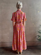 Mature woman wearing Martine maxi Dress from Trovata rendered in the Botanic Tile pink and orange floral print and crafted from a luxurious blend of cotton and silk with button front, collar, short sleeves and matching optional self tile belt