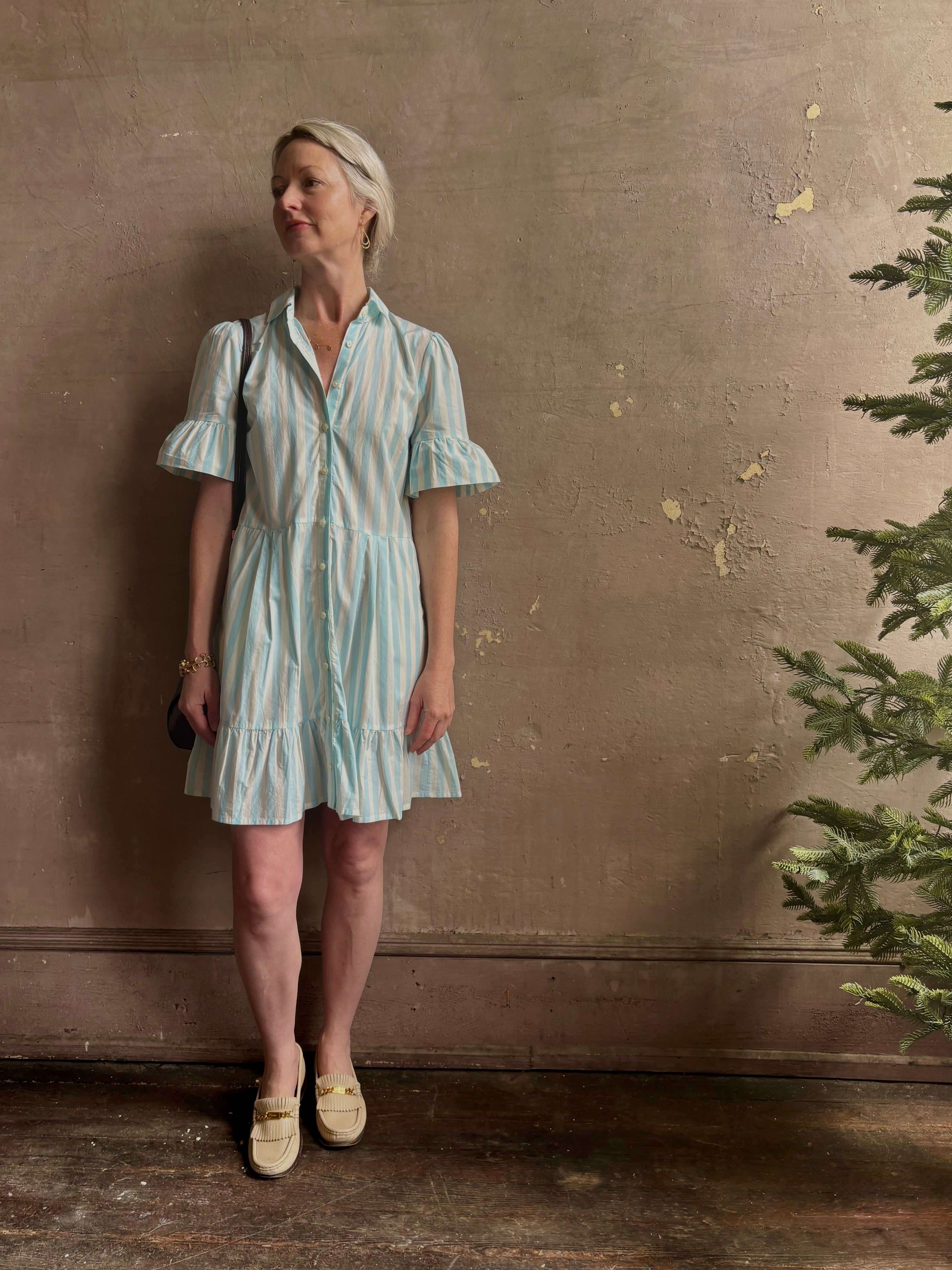 Mature woman wearing Martinique Dress from Trovata rendered in a cream and aqua Stripe and crafted from cotton poplin with ruffle trimmed short sleeves, collared button front short dress and co-ordinating fabric tie belt