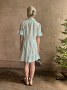 Mature woman wearing Martinique Dress from Trovata rendered in a cream and aqua Stripe and crafted from cotton poplin with ruffle trimmed short sleeves, collared button front short dress and co-ordinating fabric tie belt