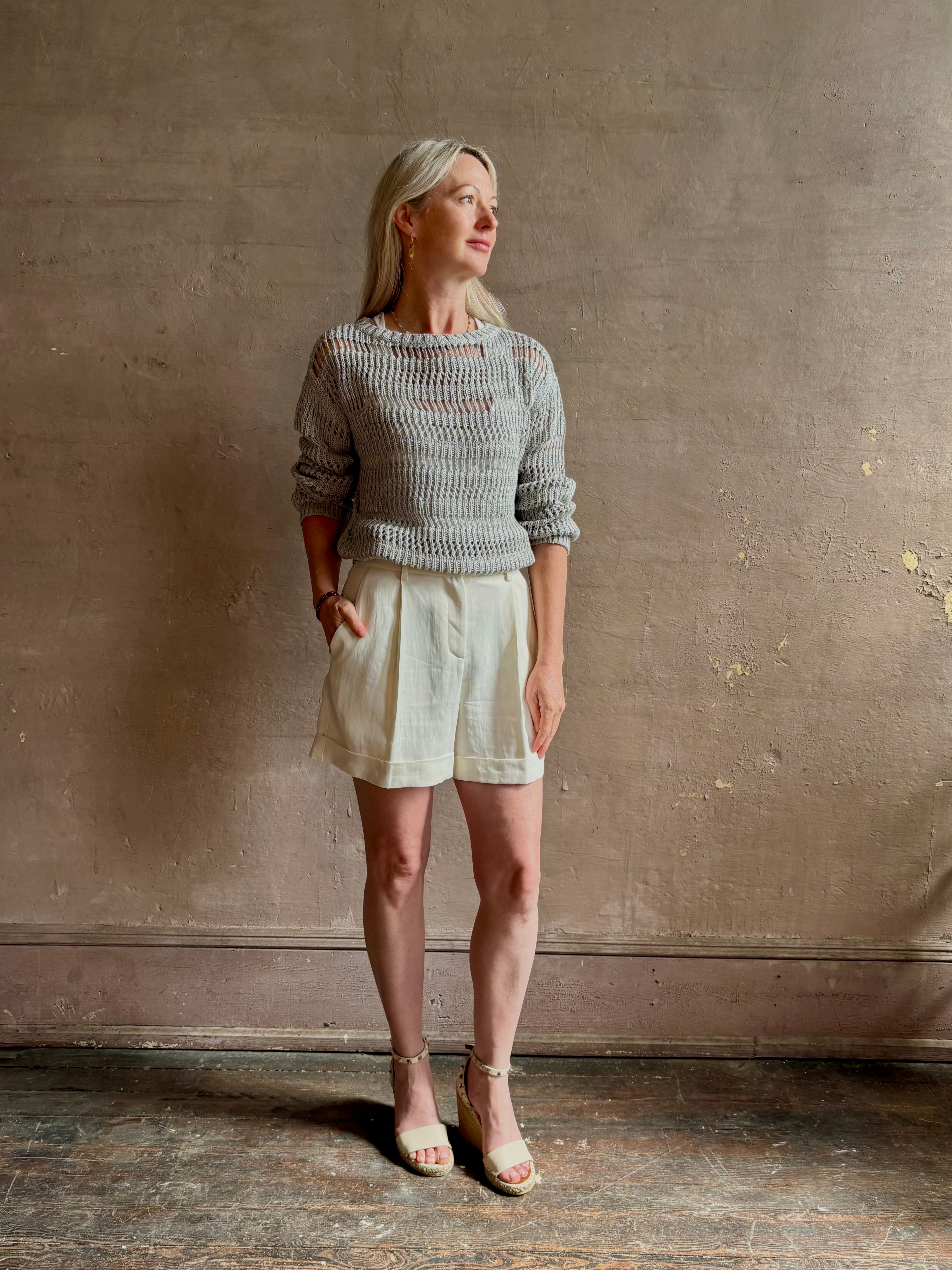 Mature woman wearing elevated tailored Cisco Shorts from TWP rendered in a soft bone white and crafted in the USA using a viscose and  linen blend fabric with side seam pockets, belt loops, single front pleat and cuffed at the bottom