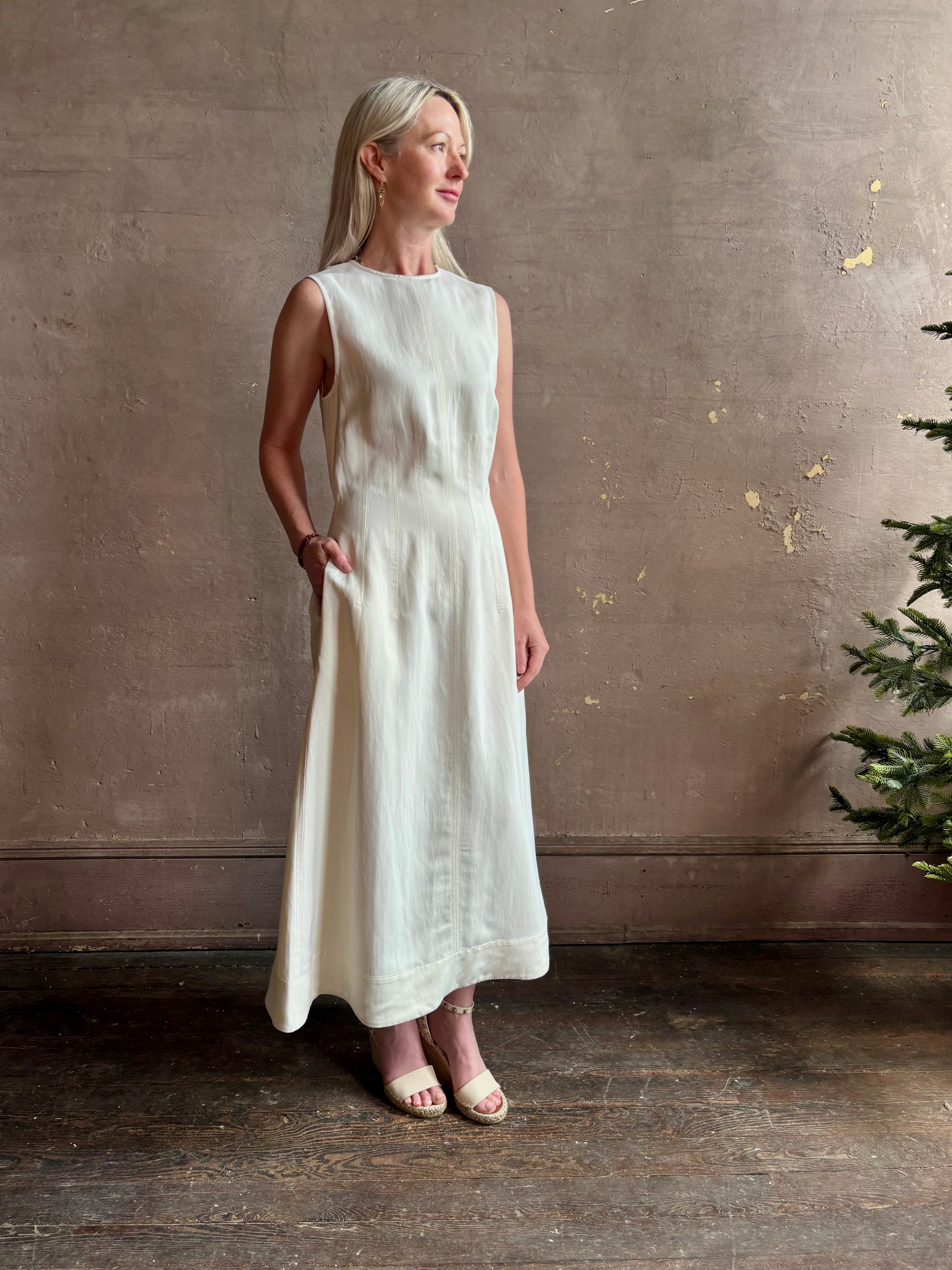 Mature woman wearing elegant Crew Neck maxi A-Line Dress with Darts from TWP rendered in a soft off white and crafted in the USA from a viscose and linen fabric blend with side seam pockets and sleeveless