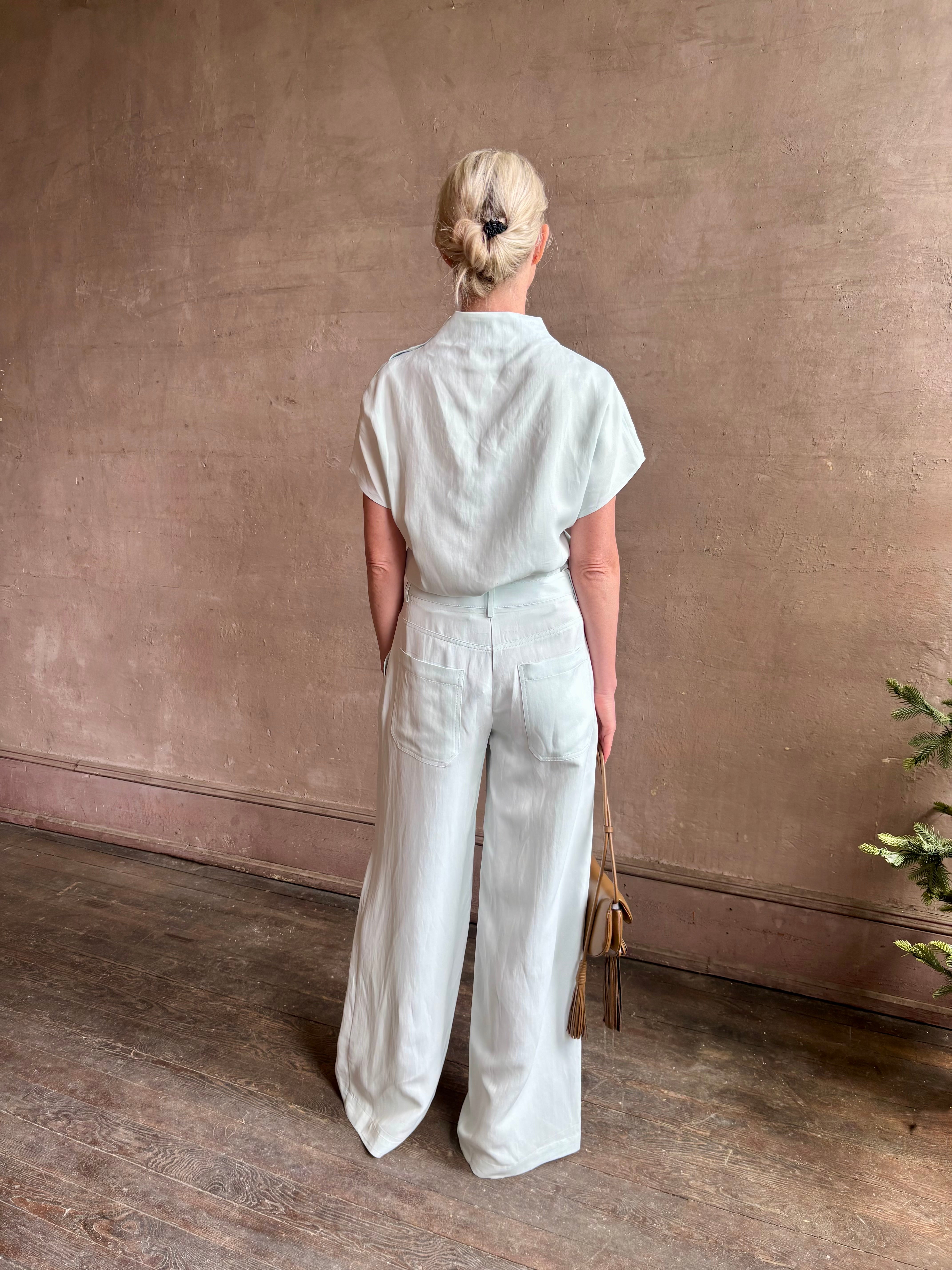 Mature woman wearing the Dylan Pant from TWP rendered in a sea glass blue hue and crafted from a viscose and linen blend, made in the USA, this luxury mid-rise trouser offers a wide leg silhouette with hidden seam side and back pockets 