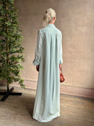 Mature woman wearing floor-length Jenny's Gown from TWP rendered in a sea glass hue and crafted from a viscose linen blend with beautiful draping, long sleeves, button front and cool girl loose fit