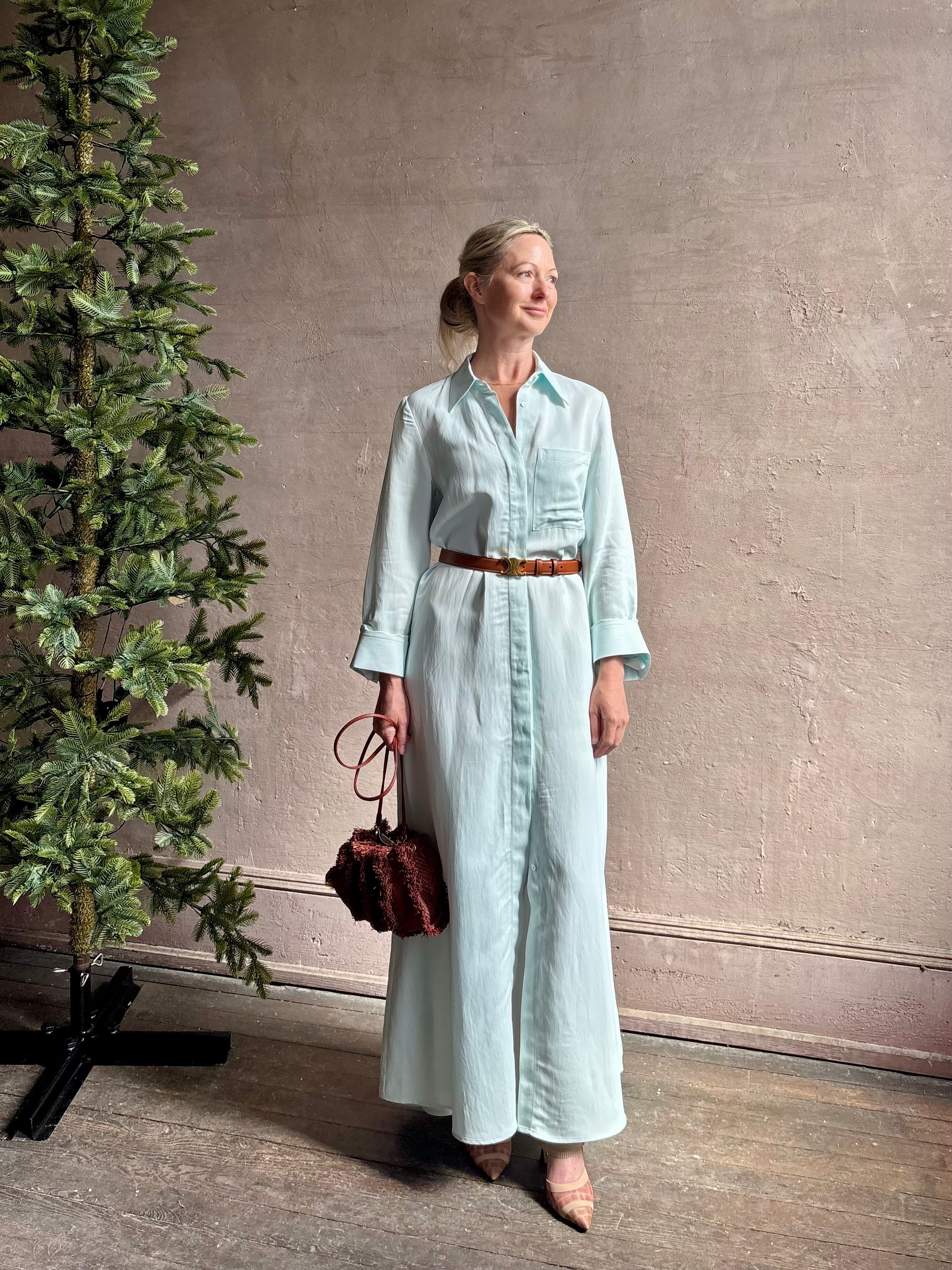 Mature woman wearing floor-length Jenny's Gown from TWP rendered in a sea glass hue and crafted from a viscose linen blend with beautiful draping, long sleeves, button front and cool girl loose fit