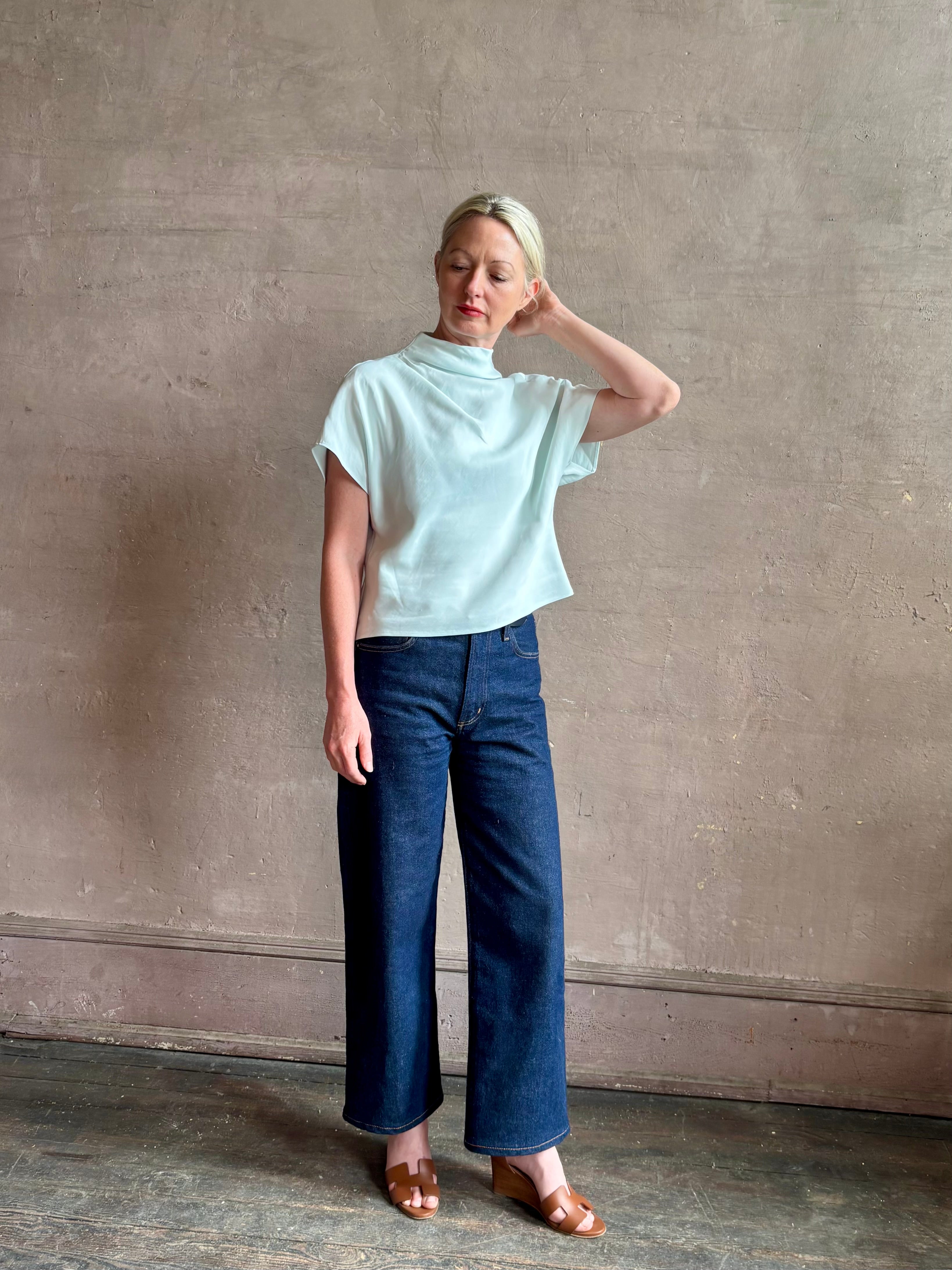 Mature woman wearing refined Mock Neck Tee Blouse from TWP rendered in a subtle sea glass blue hue and crafted from a viscose linen blend made in the USA, with short sleeves, asymmetrical design and quality fabric draping