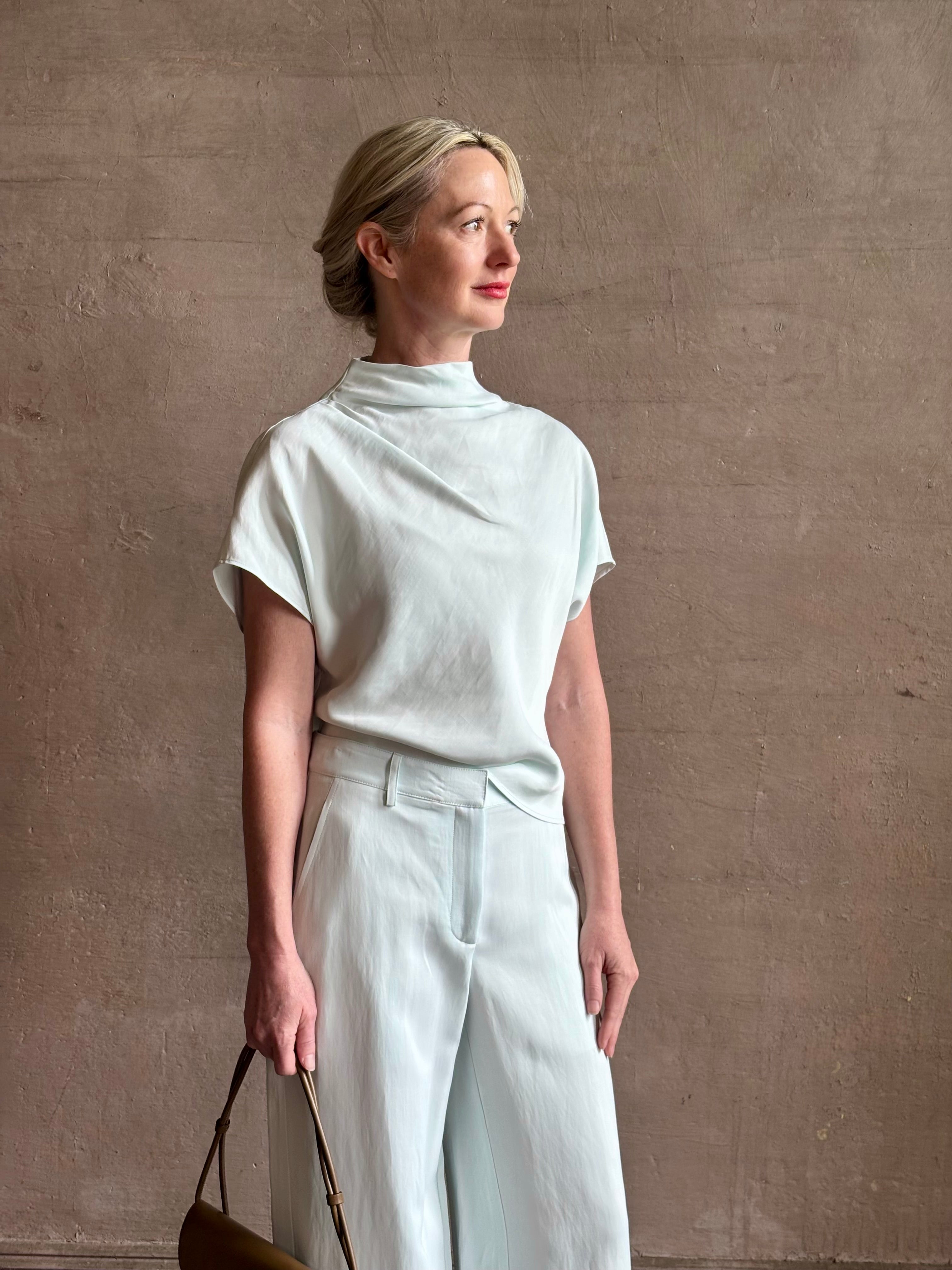 Mature woman wearing refined Mock Neck Tee Blouse from TWP rendered in a subtle sea glass blue hue and crafted from a viscose linen blend made in the USA, with short sleeves, asymmetrical design and quality fabric draping style with matching Dylan pant