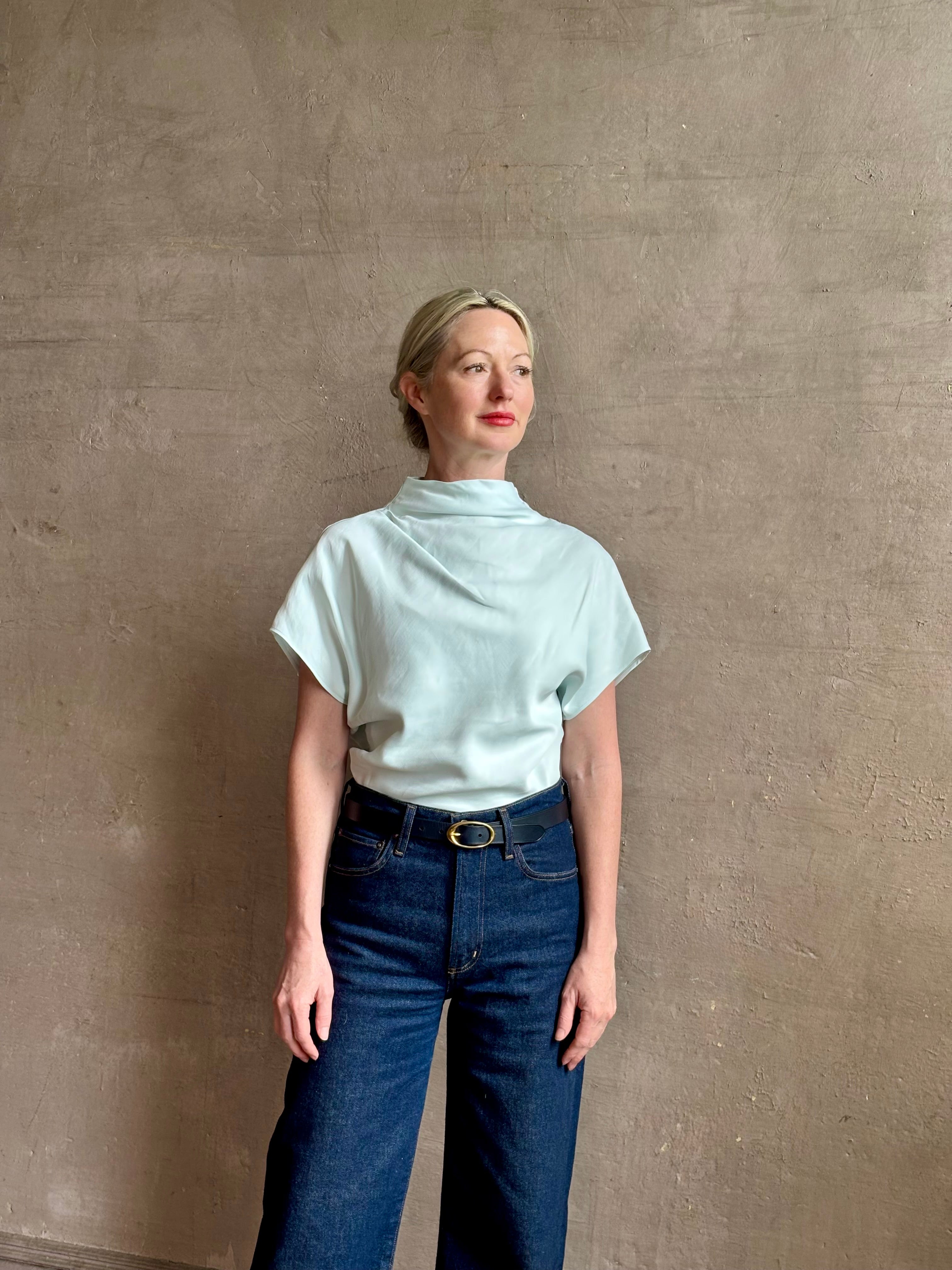 Mature woman wearing refined Mock Neck Tee Blouse from TWP rendered in a subtle sea glass blue hue and crafted from a viscose linen blend made in the USA, with short sleeves, asymmetrical design and quality fabric draping