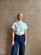 Mature woman wearing refined Mock Neck Tee Blouse from TWP rendered in a subtle sea glass blue hue and crafted from a viscose linen blend made in the USA, with short sleeves, asymmetrical design and quality fabric draping