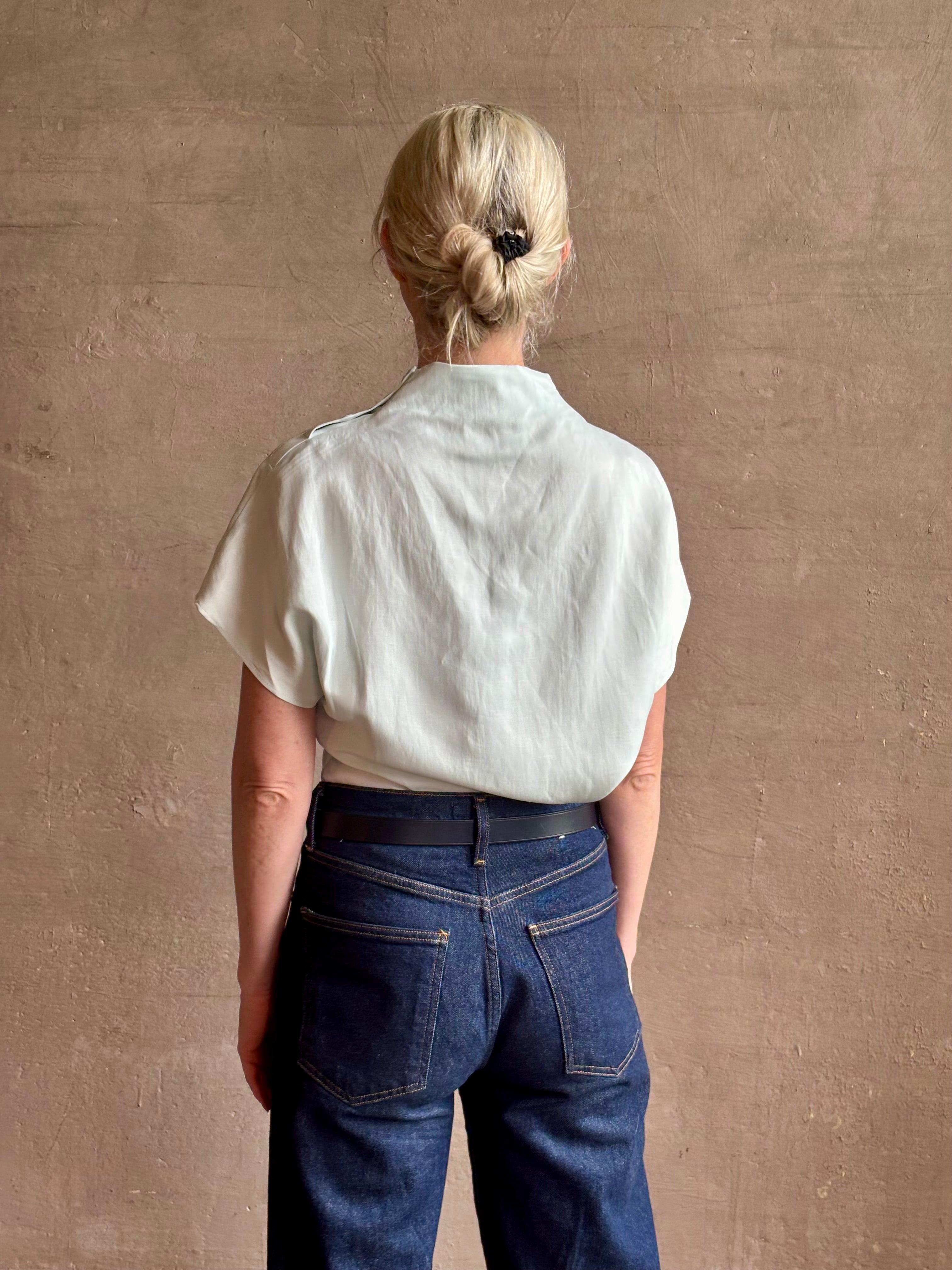 Mature woman wearing refined Mock Neck Tee Blouse from TWP rendered in a subtle sea glass blue hue and crafted from a viscose linen blend made in the USA, with short sleeves, asymmetrical design and quality fabric draping