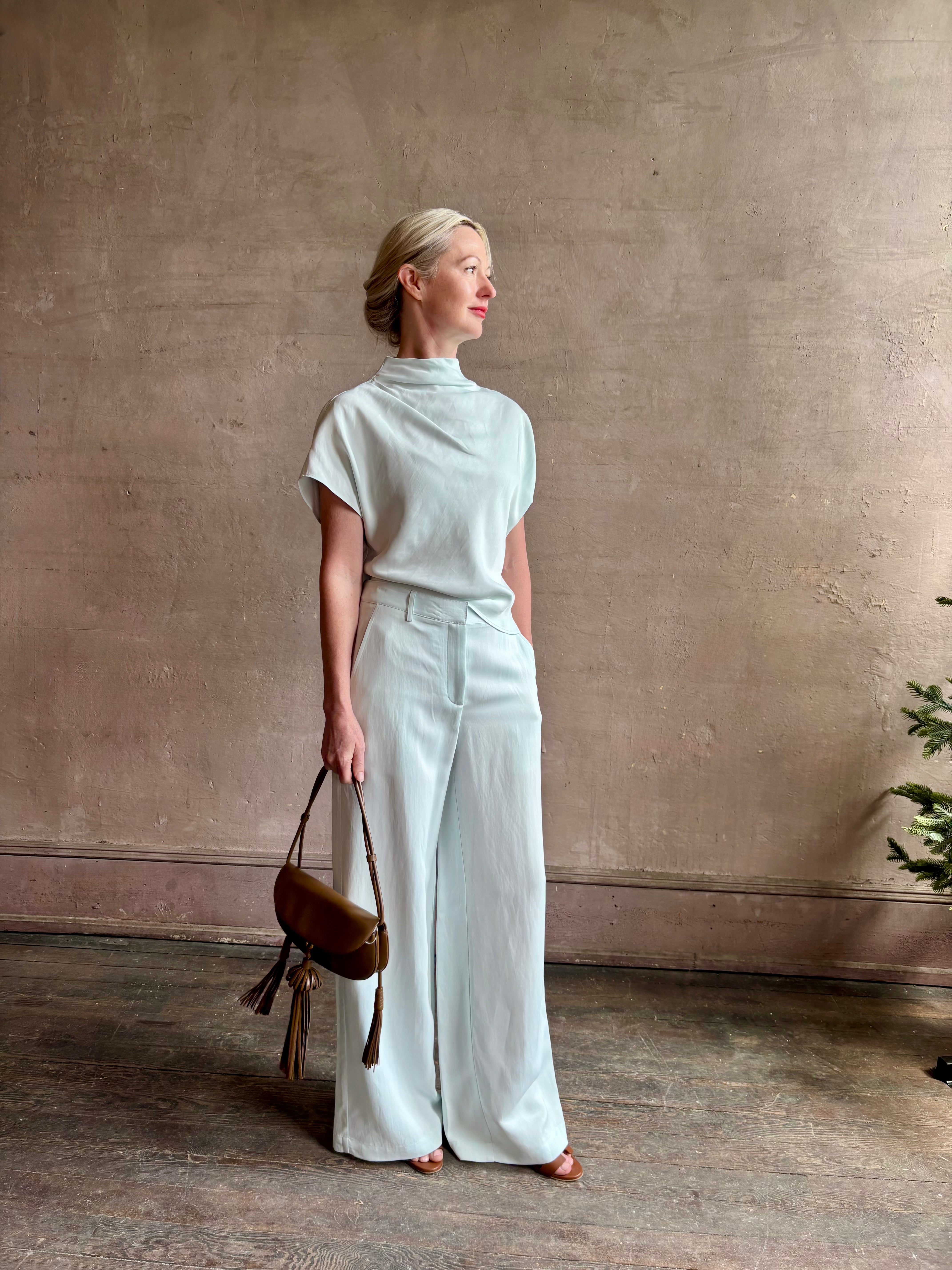 Mature woman wearing refined Mock Neck Tee Blouse from TWP rendered in a subtle sea glass blue hue and crafted from a viscose linen blend made in the USA, with short sleeves, asymmetrical design and quality fabric draping