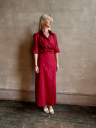 Mature woman wearing New Earl wrap shirt Dress from TWP rendered in a vibrant red and crafted from cotton poplin in the USA with 1/2 sleeves, collar, asymetrical button top and maxi length dress