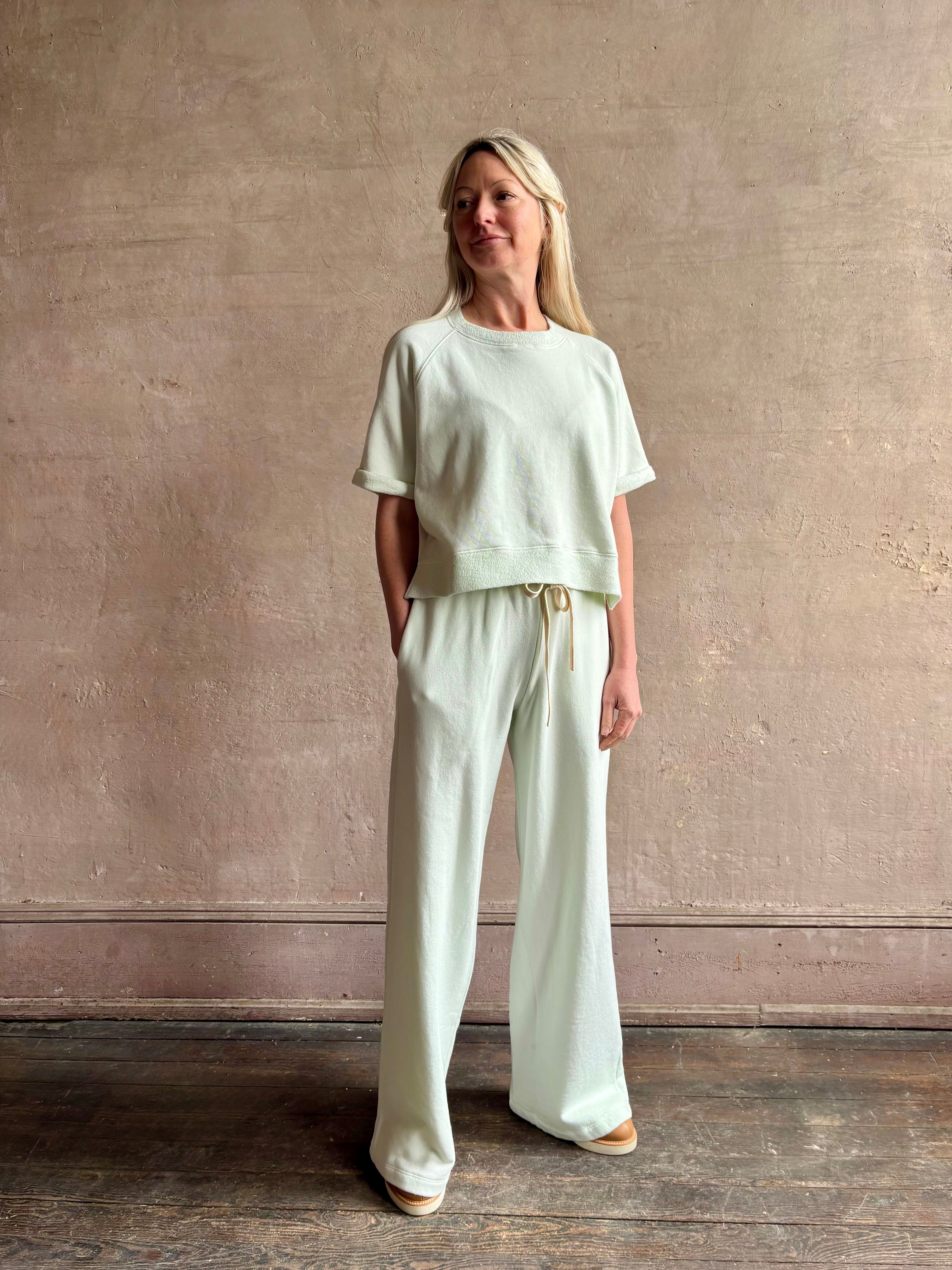 Mature woman wearing Slouchy Rolled Tee Top from TWP rendered in a sea glass hue and crafted from cotton with raglan short sleeves, crew neck and relaxed silhouette styled with matching PJ pant for the complete luxury lounge set