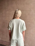 Mature woman wearing Slouchy Rolled Tee Top from TWP rendered in a sea glass hue and crafted from cotton with raglan short sleeves, crew neck and relaxed silhouette