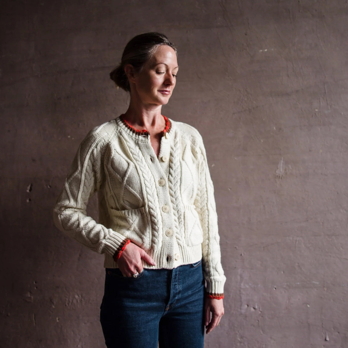 Image featuring a woman wearing the Louise classic cable knit button down cardigan from Trovata with red and brown finishing trim edges.