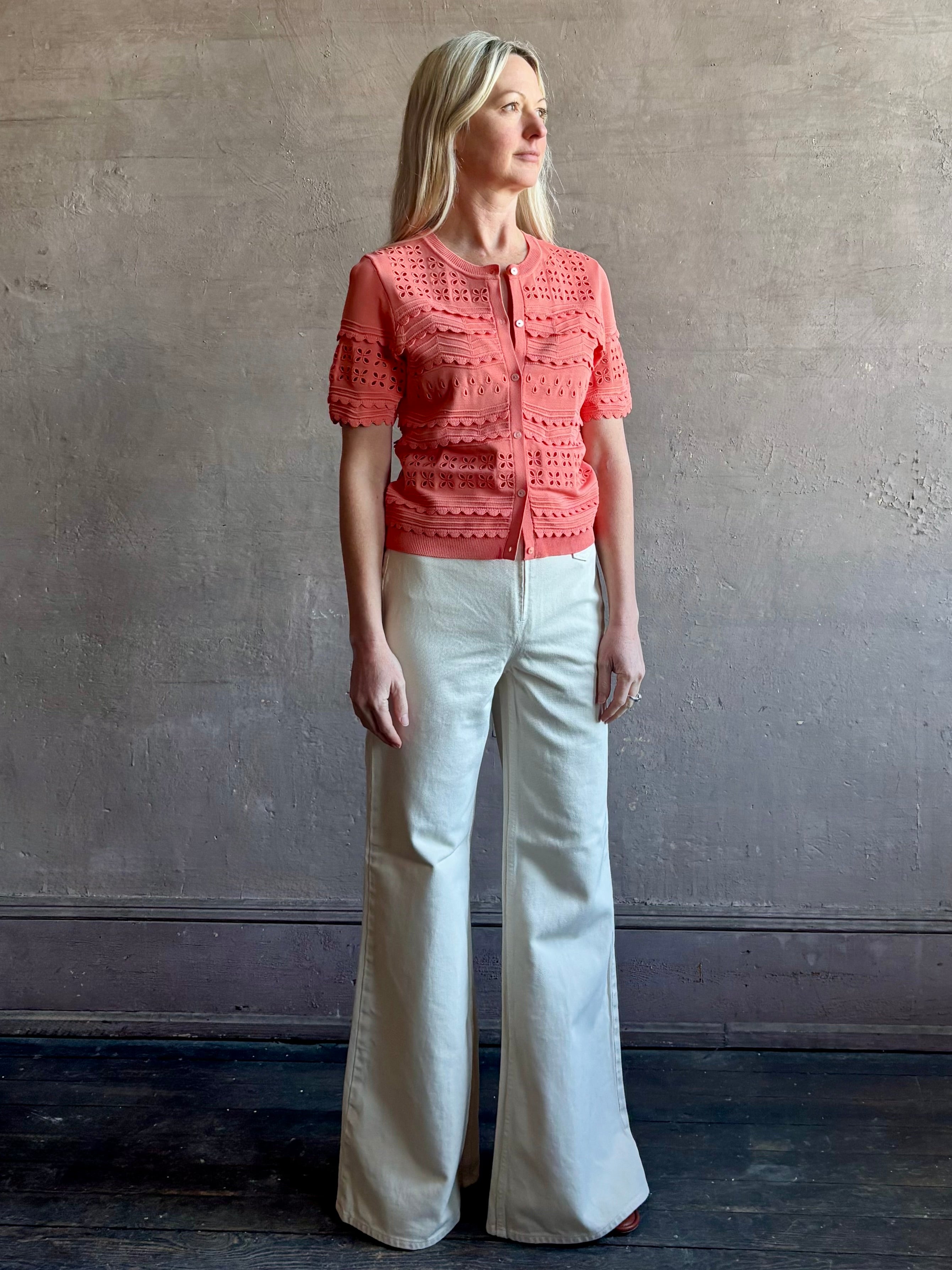 Woman wearing the romantic Abriana Eyelet Knit Cardigan from Ulla Johnson rendered in a luminous coral hue and crafted from luxury knit with round neck, button front and short sleeves