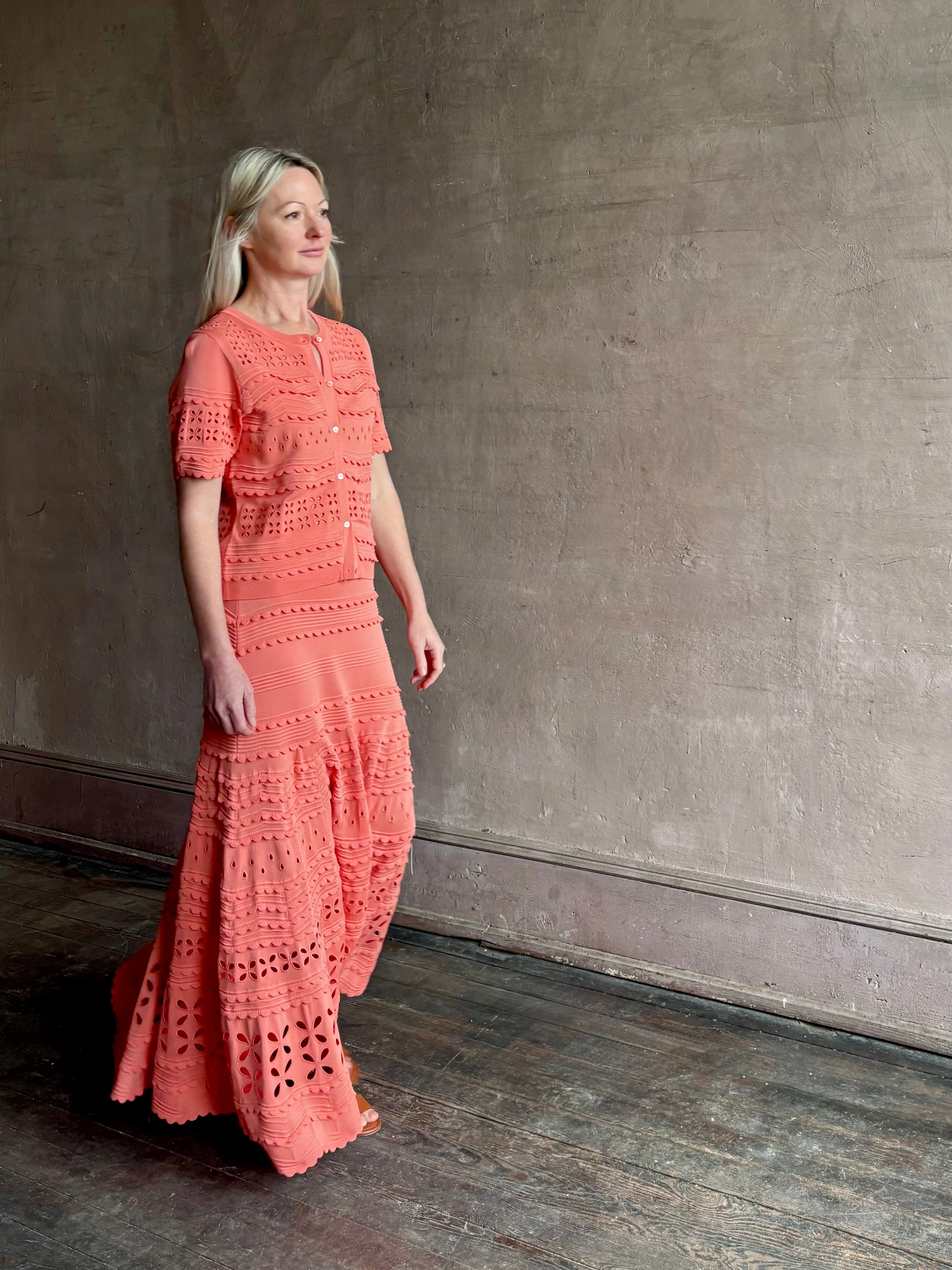 Woman wearing the feminine  Annisa Eyelet Knit A-line maxi skirt from Ulla Johnson rendered in a luminous coral hue and crafted from luxury knit with matching top