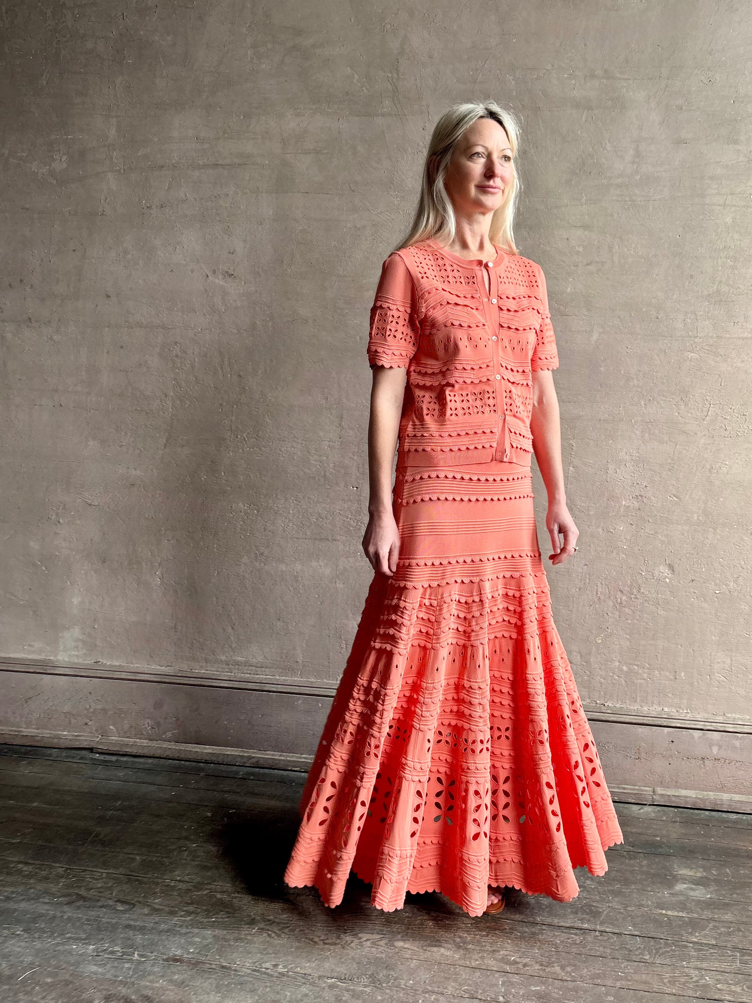 Woman wearing the feminine  Annisa Eyelet Knit A-line maxi skirt from Ulla Johnson rendered in a luminous coral hue and crafted from luxury knit with matching top