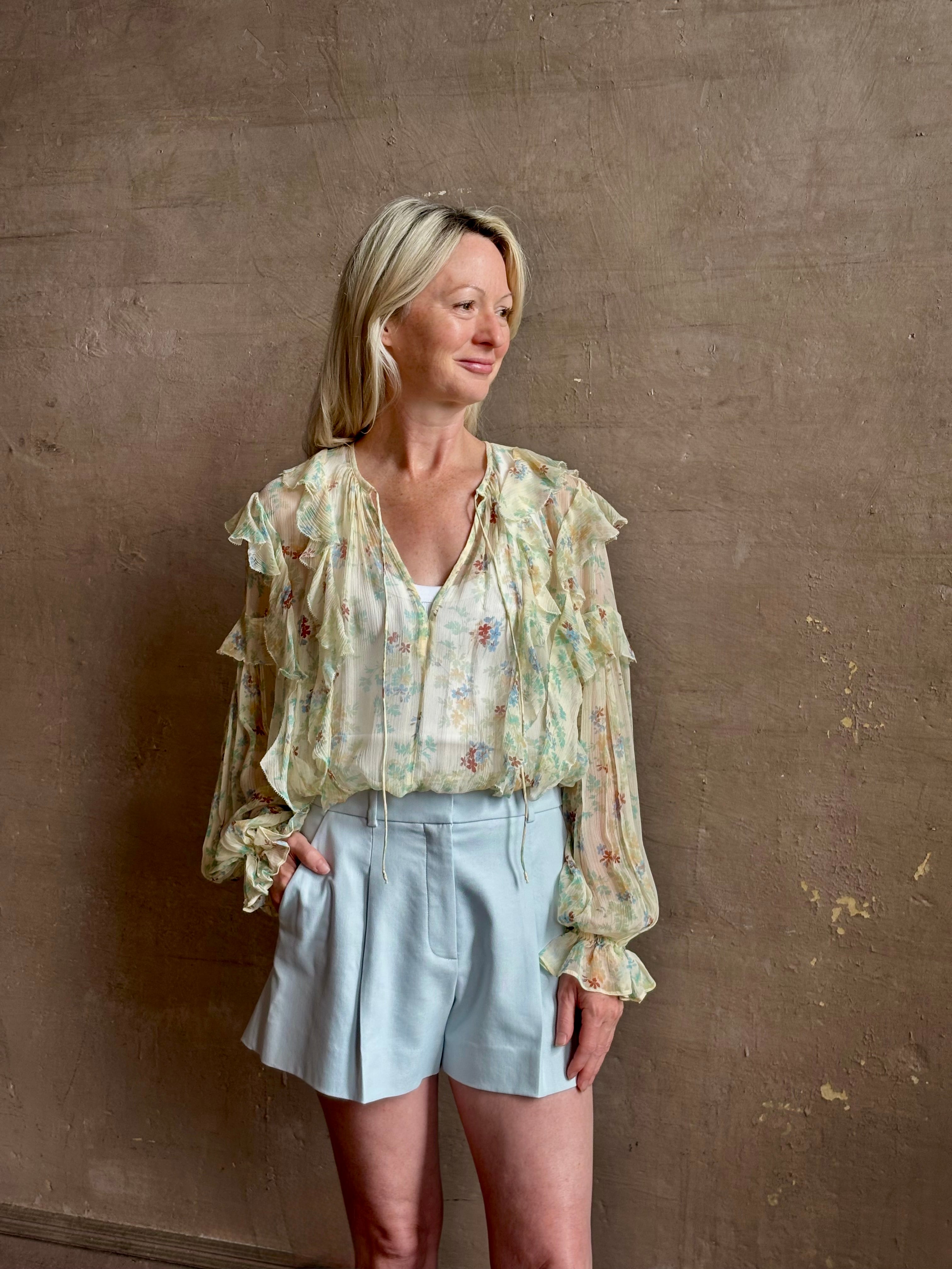 Mature woman wearing Delfina Long Sleeve Silk Blouse from Ulla Johnson rendered in a light yellow body adorned with a delicate, scattered tiny floral print with v-neck, drawstring waist, ruffle lined sleeves and linear ruffle details at front and back