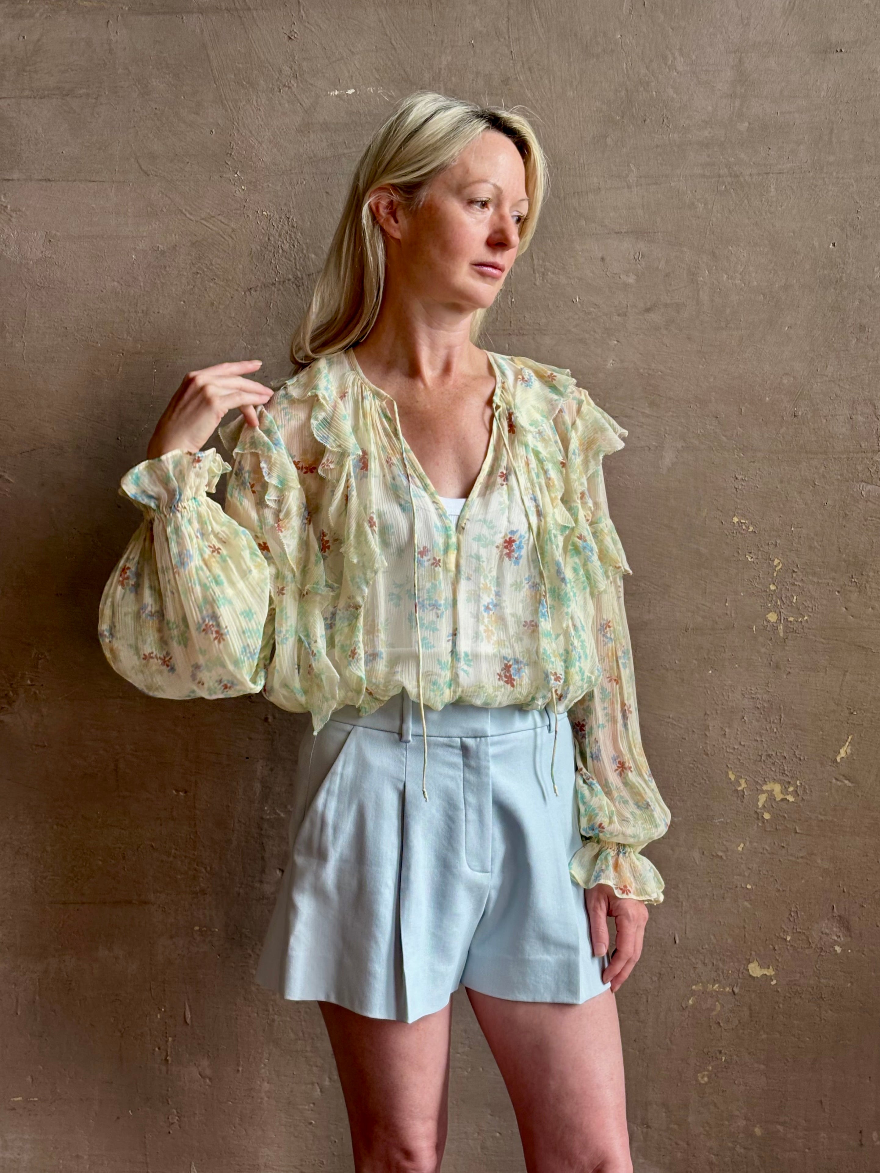 Mature woman wearing Delfina Long Sleeve Silk Blouse from Ulla Johnson rendered in a light yellow body adorned with a delicate, scattered tiny floral print with v-neck, drawstring waist, ruffle lined sleeves and linear ruffle details at front and back