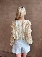 Mature woman wearing Delfina Long Sleeve Silk Blouse from Ulla Johnson rendered in a light yellow body adorned with a delicate, scattered tiny floral print with v-neck, drawstring waist, ruffle lined sleeves and linear ruffle details at front and back