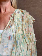 Mature woman wearing Delfina Long Sleeve Silk Blouse from Ulla Johnson rendered in a light yellow body adorned with a delicate, scattered tiny floral print with v-neck, drawstring waist, ruffle lined sleeves and linear ruffle details at front and back