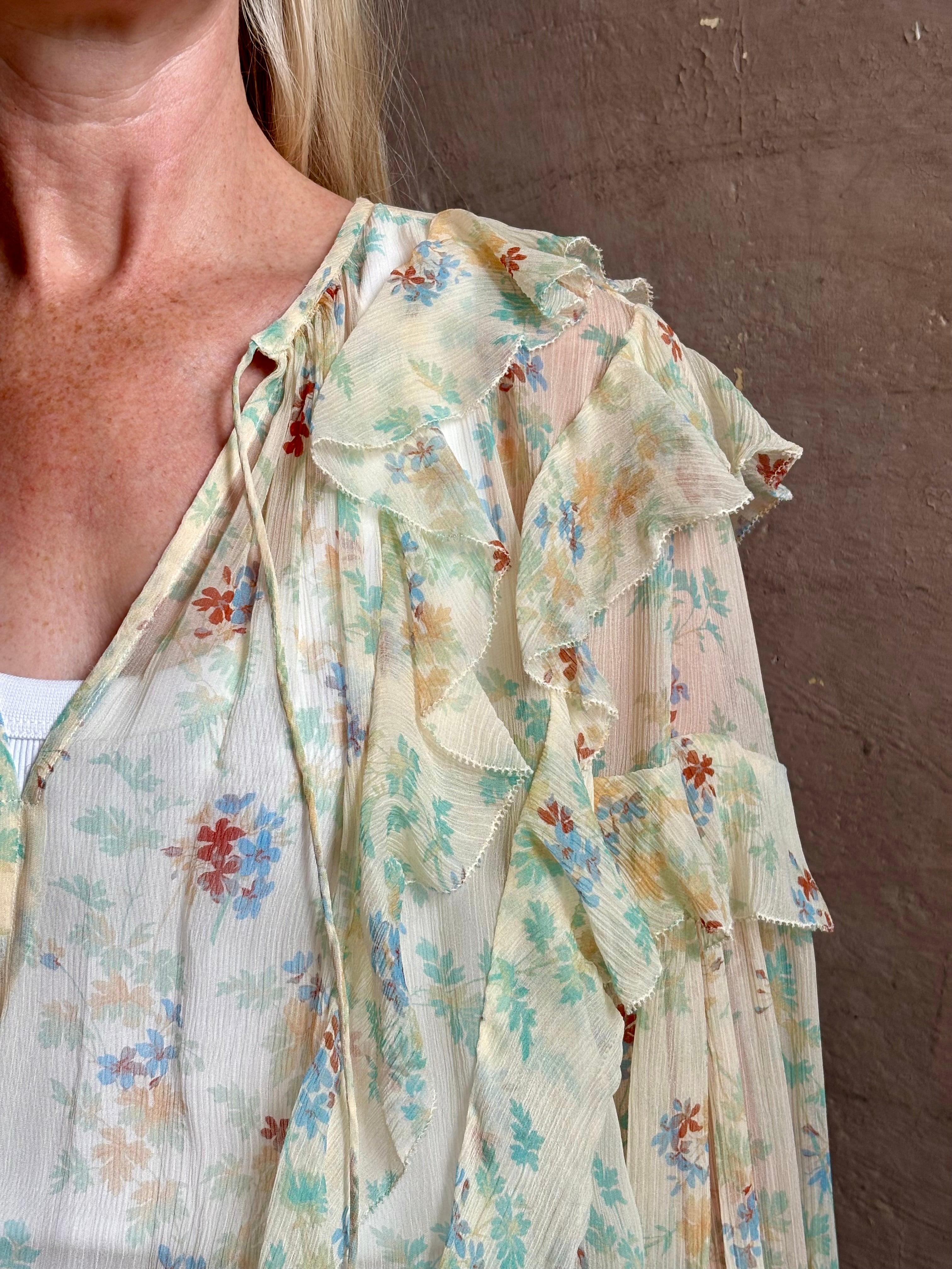 Mature woman wearing Delfina Long Sleeve Silk Blouse from Ulla Johnson rendered in a light yellow body adorned with a delicate, scattered tiny floral print with v-neck, drawstring waist, ruffle lined sleeves and linear ruffle details at front and back