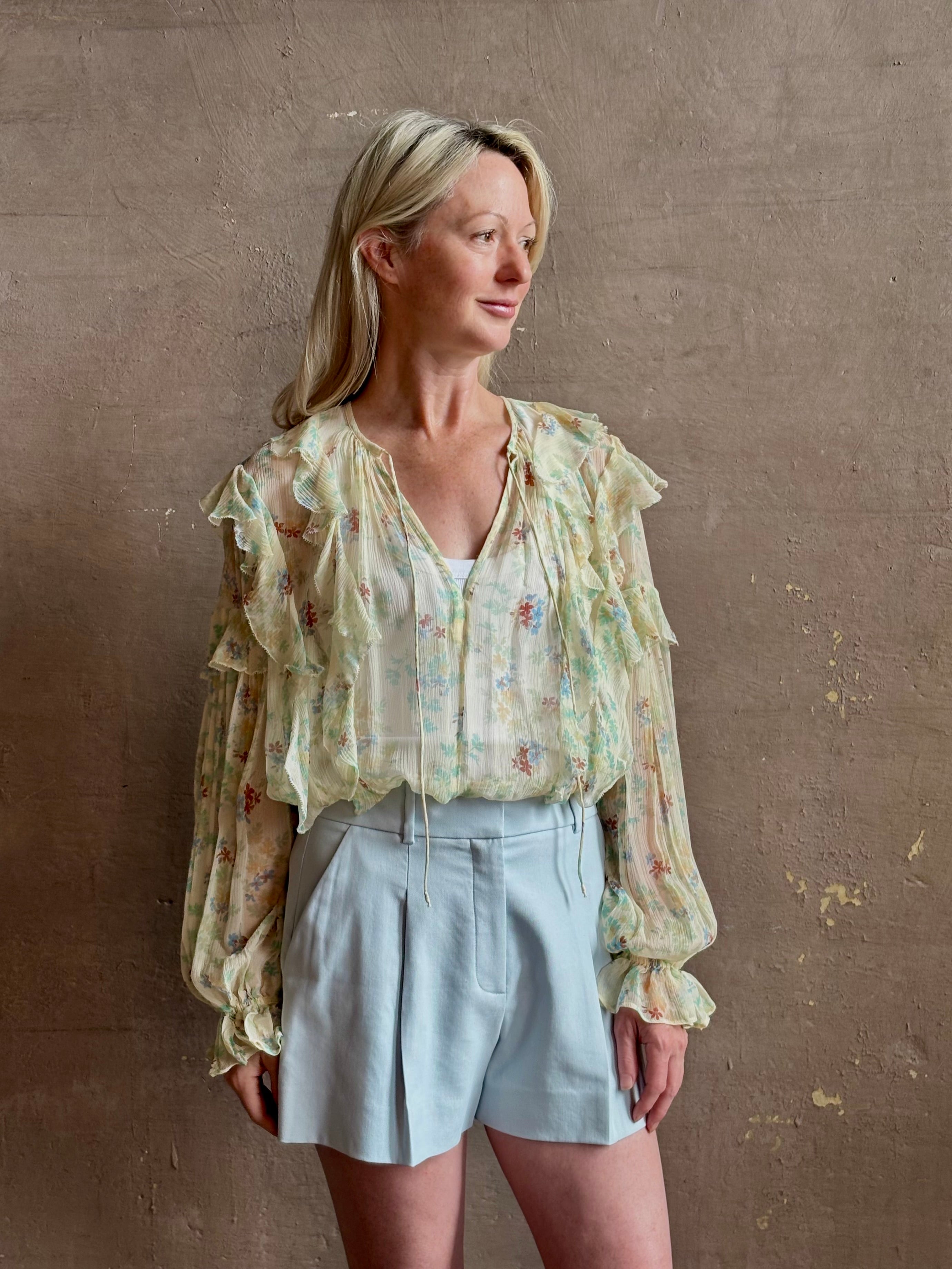 Mature woman wearing Delfina Long Sleeve Silk Blouse from Ulla Johnson rendered in a light yellow body adorned with a delicate, scattered tiny floral print with v-neck, drawstring waist, ruffle lined sleeves and linear ruffle details at front and back