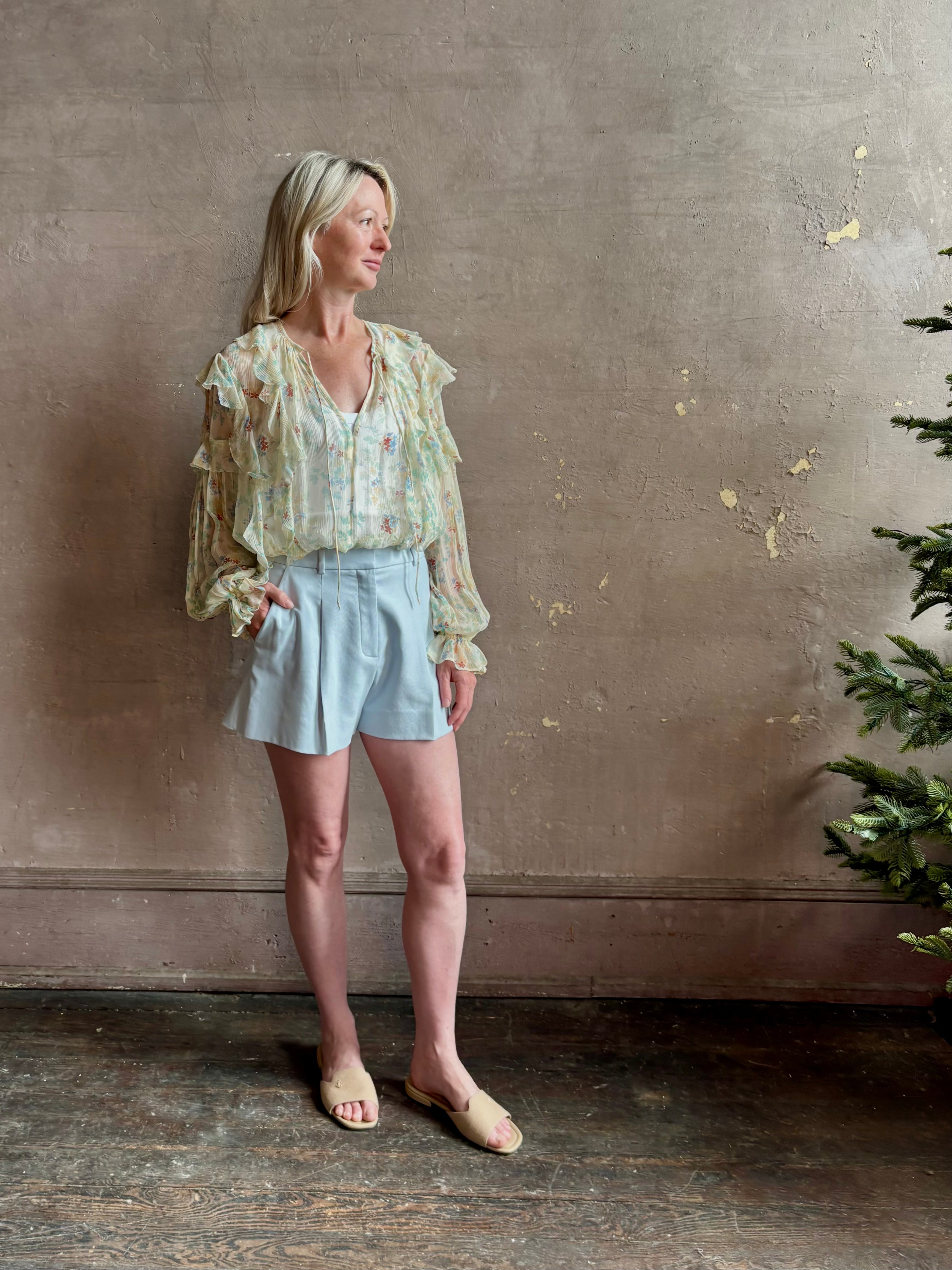 Mature woman wearing Delfina Long Sleeve Silk Blouse from Ulla Johnson rendered in a light yellow body adorned with a delicate, scattered tiny floral print with v-neck, drawstring waist, ruffle lined sleeves and linear ruffle details at front and back