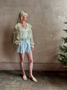 Mature woman wearing Delfina Long Sleeve Silk Blouse from Ulla Johnson rendered in a light yellow body adorned with a delicate, scattered tiny floral print with v-neck, drawstring waist, ruffle lined sleeves and linear ruffle details at front and back