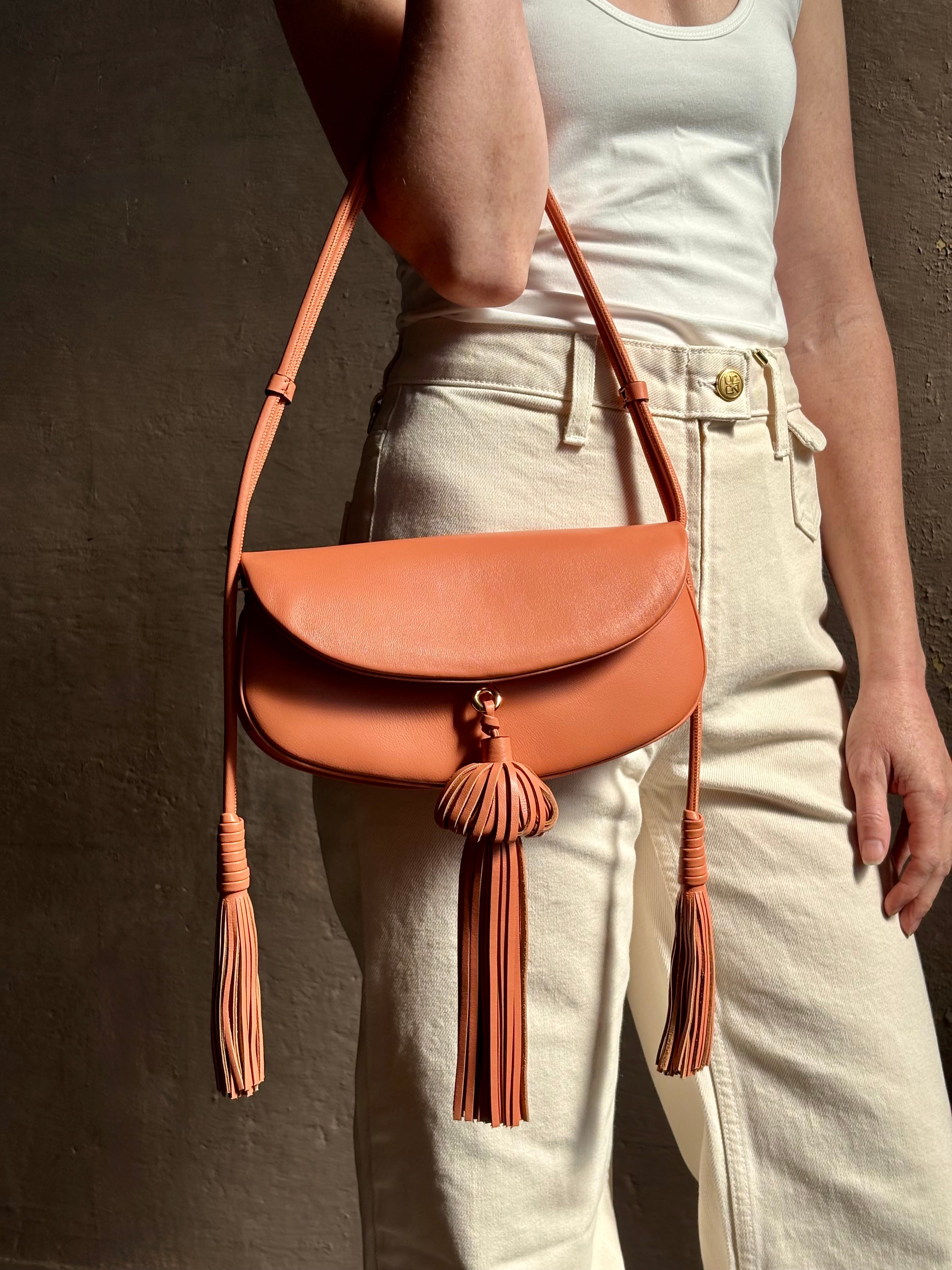 Mature woman with the luxury Italian leather  Paloma handbag with Tassel Flap from Ulla Johnson rendered in a warm pink featuring a pull tassel magnetic snap closure and removable adjustable handle that seamlessly converts to a designer clutch