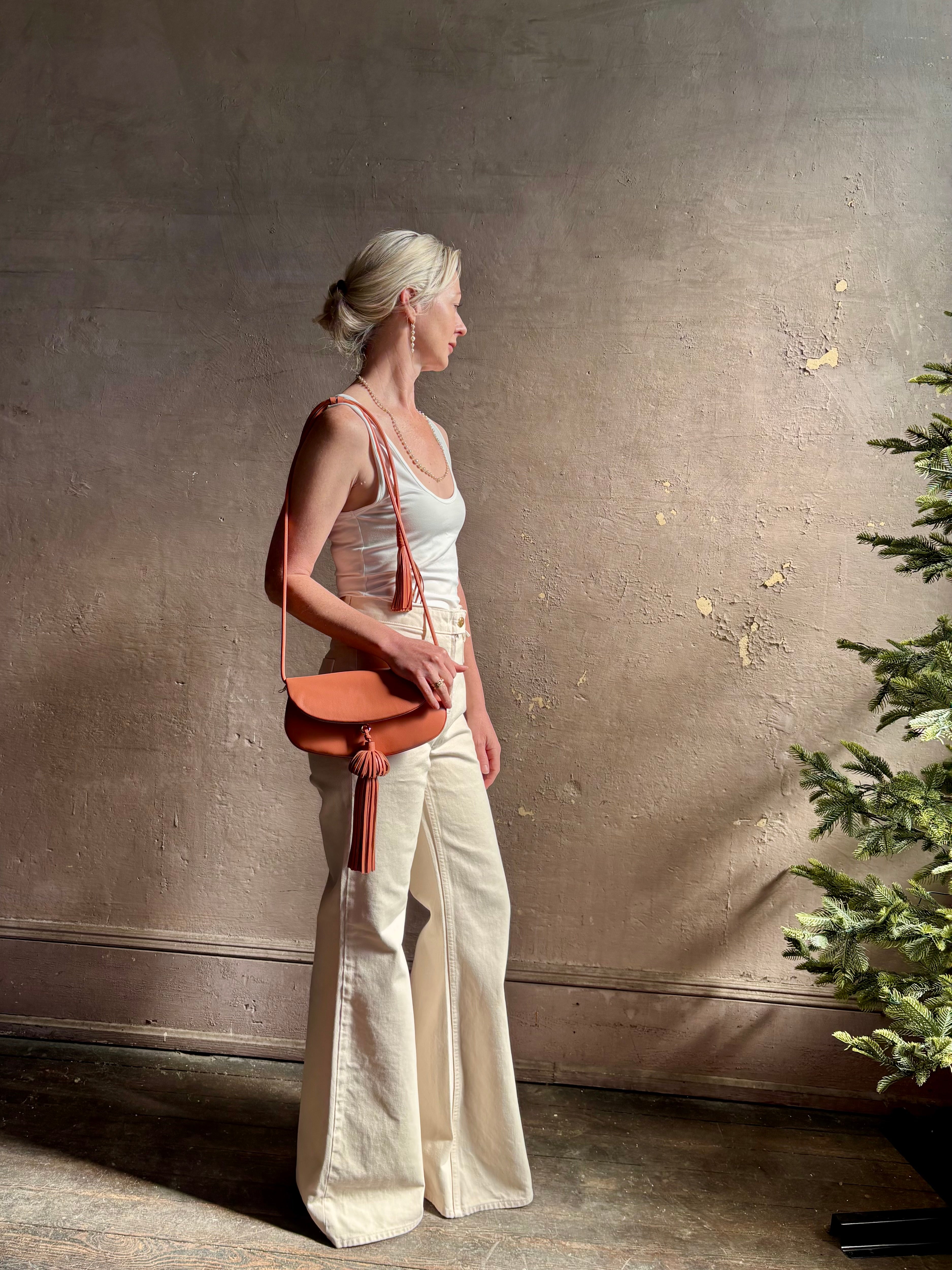 Mature woman with the luxury Italian leather  Paloma handbag with Tassel Flap from Ulla Johnson rendered in a warm pink featuring a pull tassel magnetic snap closure and removable adjustable handle that seamlessly converts to a designer clutch