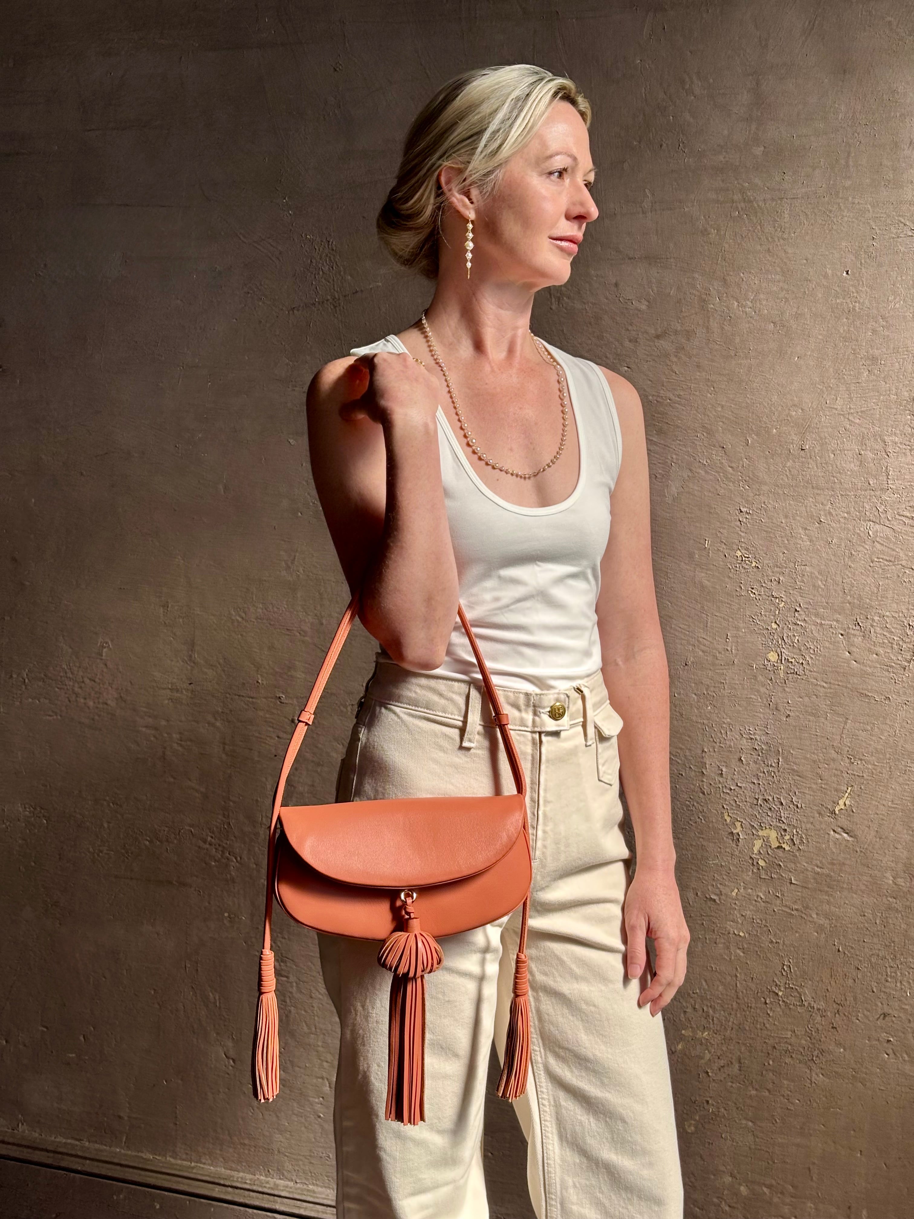 Mature woman with the luxury Italian leather  Paloma handbag with Tassel Flap from Ulla Johnson rendered in a warm pink featuring a pull tassel magnetic snap closure and removable adjustable handle that seamlessly converts to a designer clutch