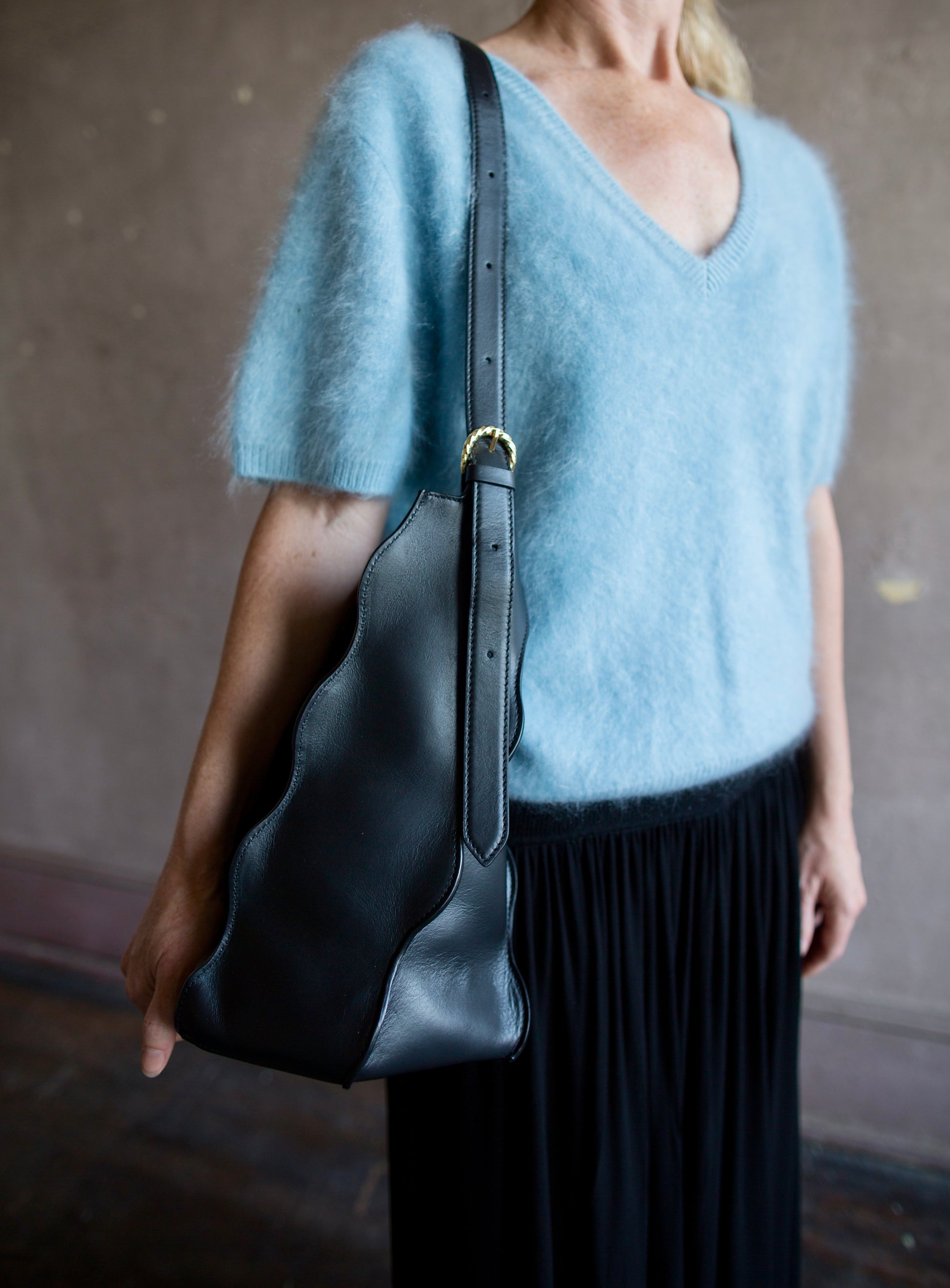 Image of a woman wearing the Adria Wave handbag by Ulla Johnson with curved seam detailing and a detachable snap pouch and adjustable strap.