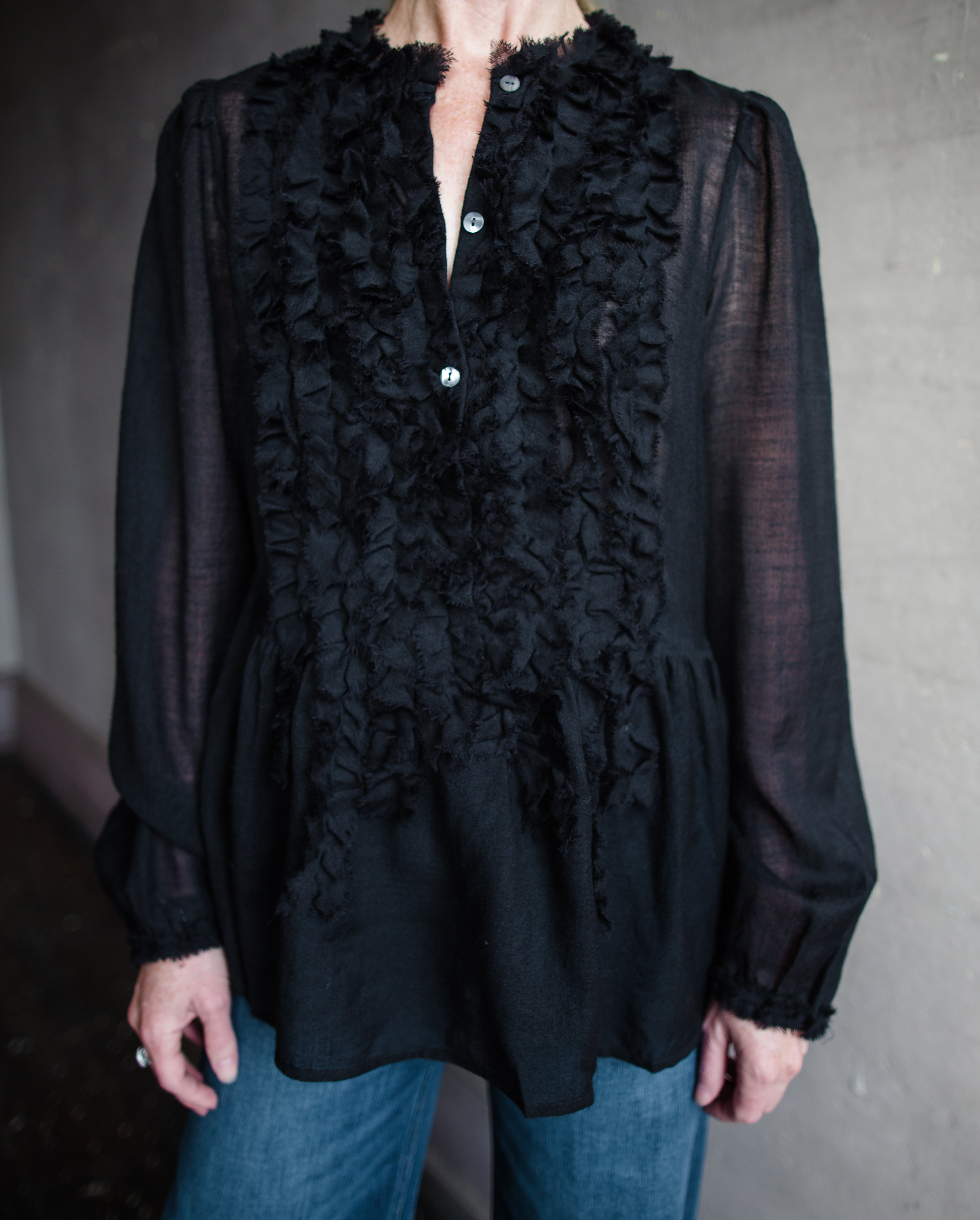 Image features woman wearing The Cosima Tunic by Ulla Johnson in Noir features a button down v neckline with rows of ruffles and iridescent sea shell buttons.